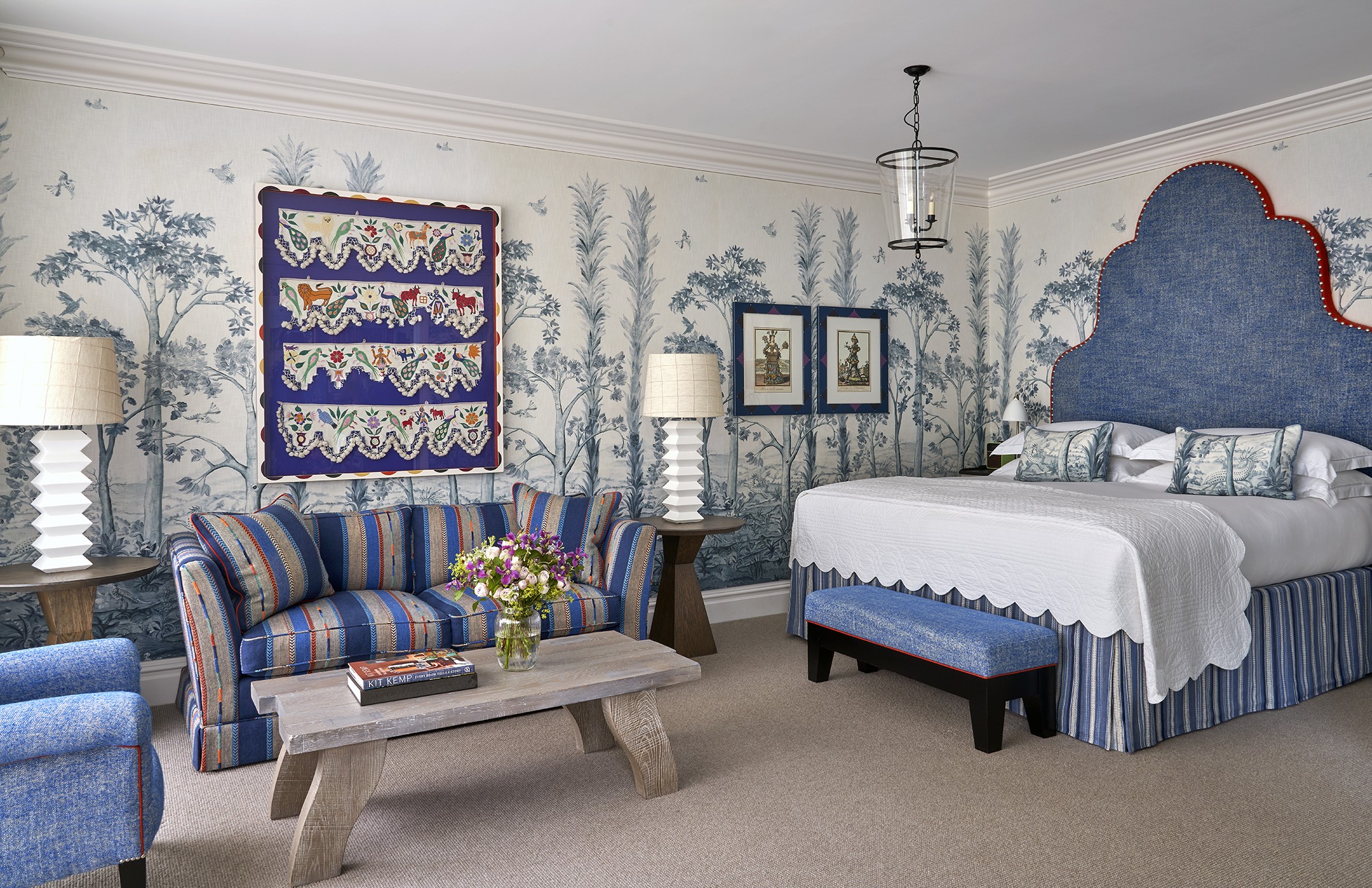 A diagonal shot of the bedroom in a Deluxe Junior Snug Suite at Ham Yard Hotel, featuring a king bed with a large headboard, a blue sofa, armchair and coffee table with two small side tables and lamps either side