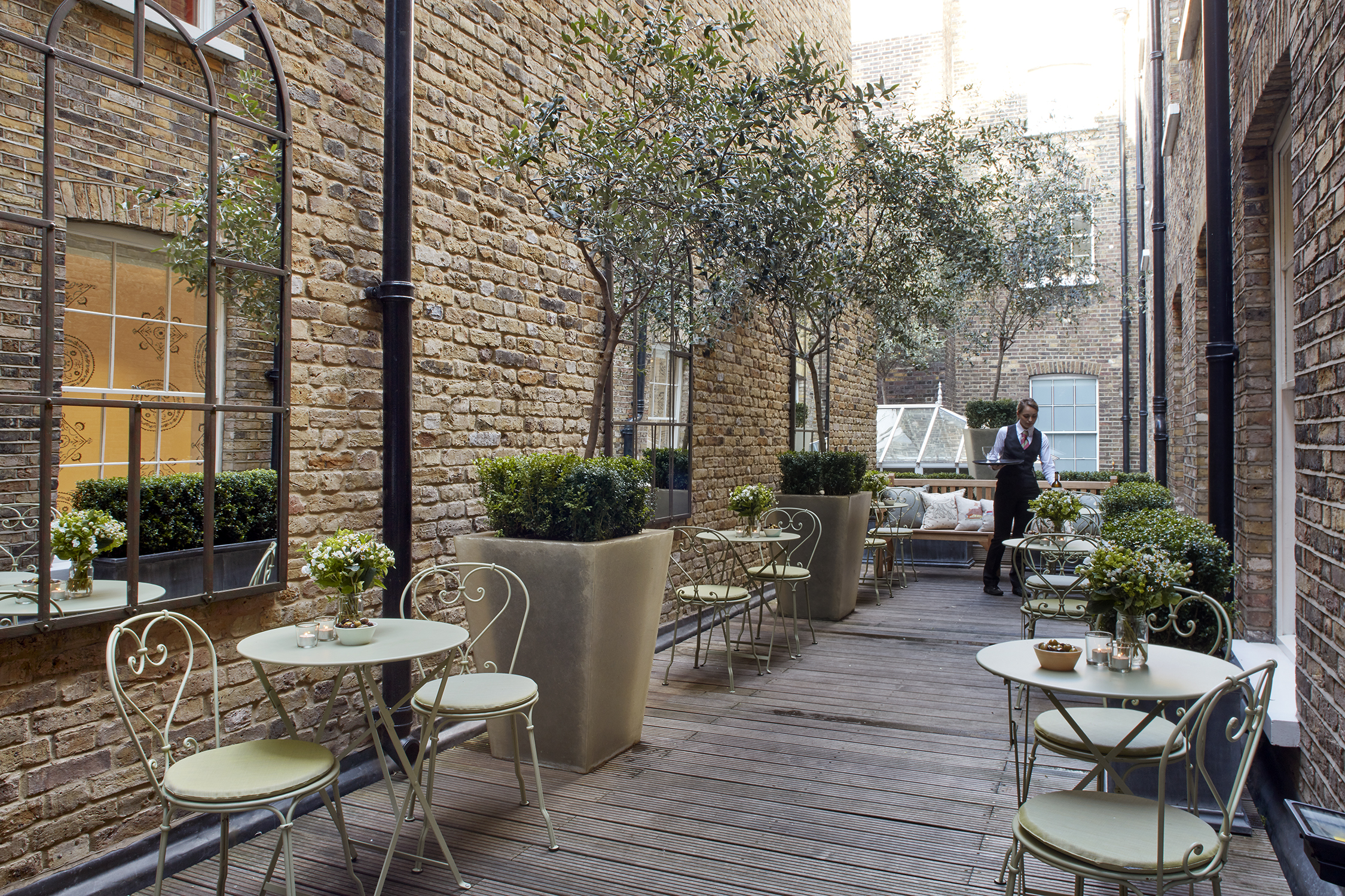 A narrow terrace lined with tables of two on the first floor at Haymarket Hotel. A waitress holds a tray at the far end