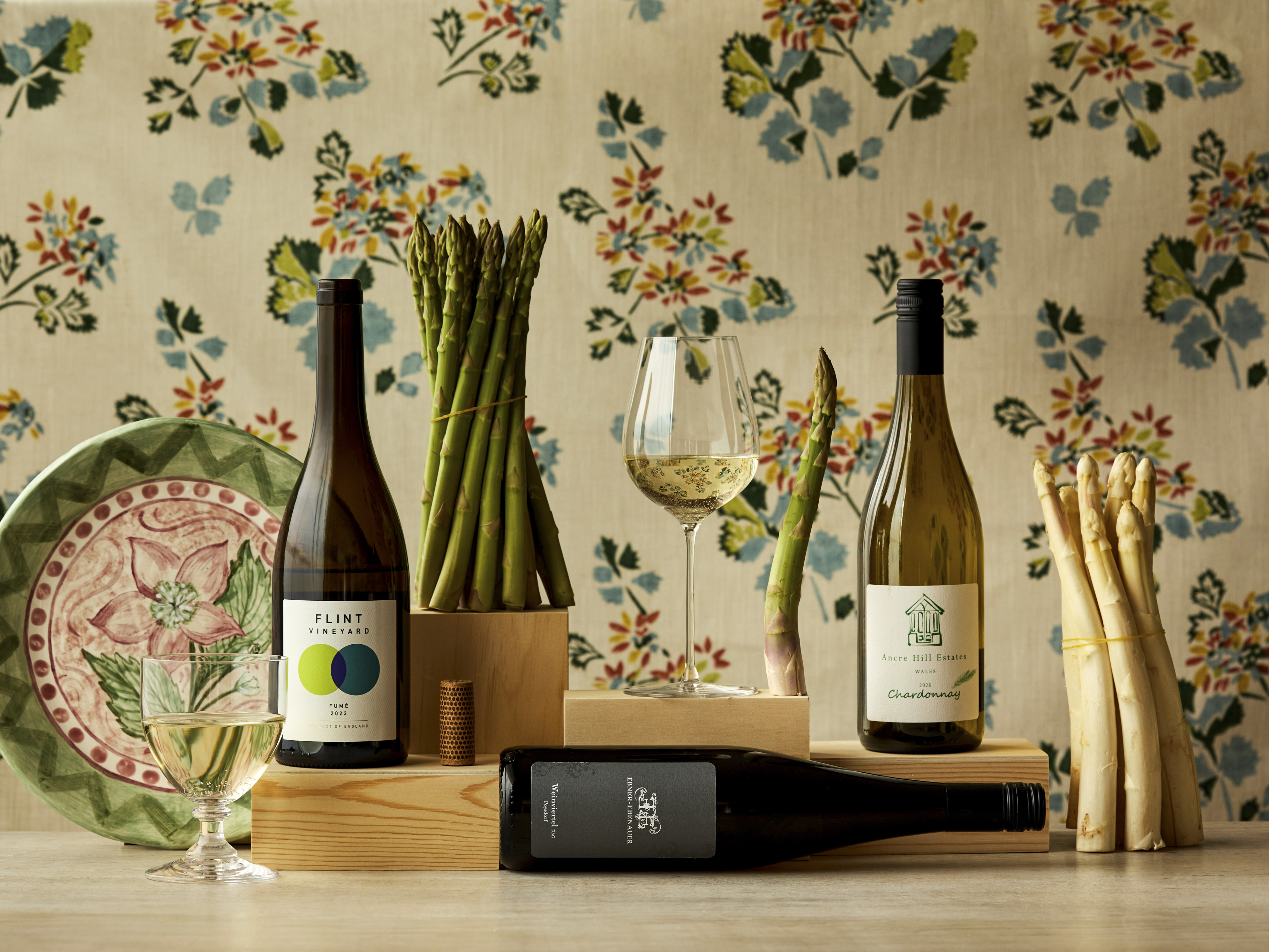 Wide shot of asparagus with wine bottles, glasses and a hand-painted ceramic plate set against and floral fabric backdrop