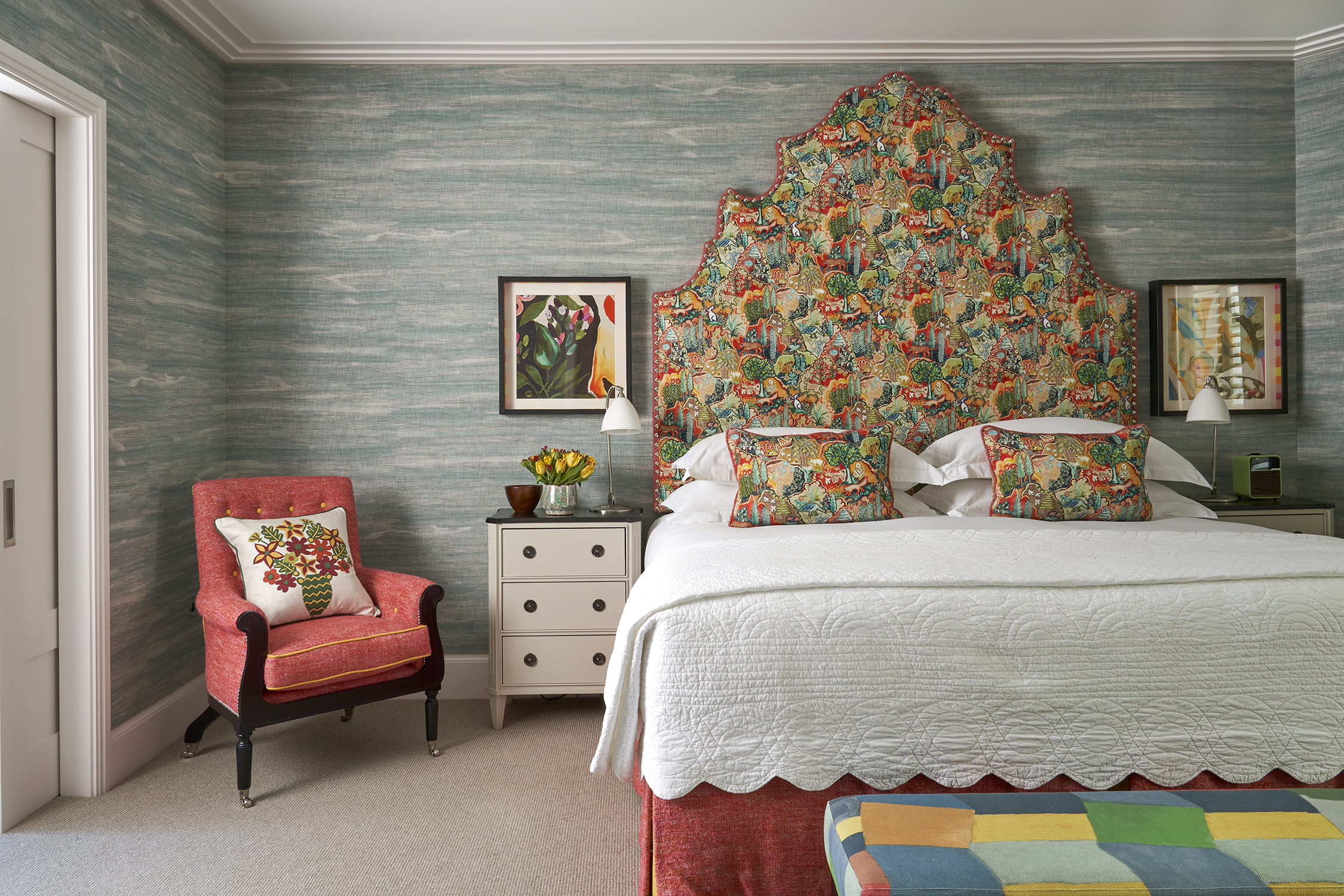 A front on image of the bedroom of The Deluxe Two Bedroom Suite, Room 5. A stylish bedroom featuring a vibrant patterned headboard and pillows, blue textured wallpaper, eclectic artwork, and an arm chair in the room.