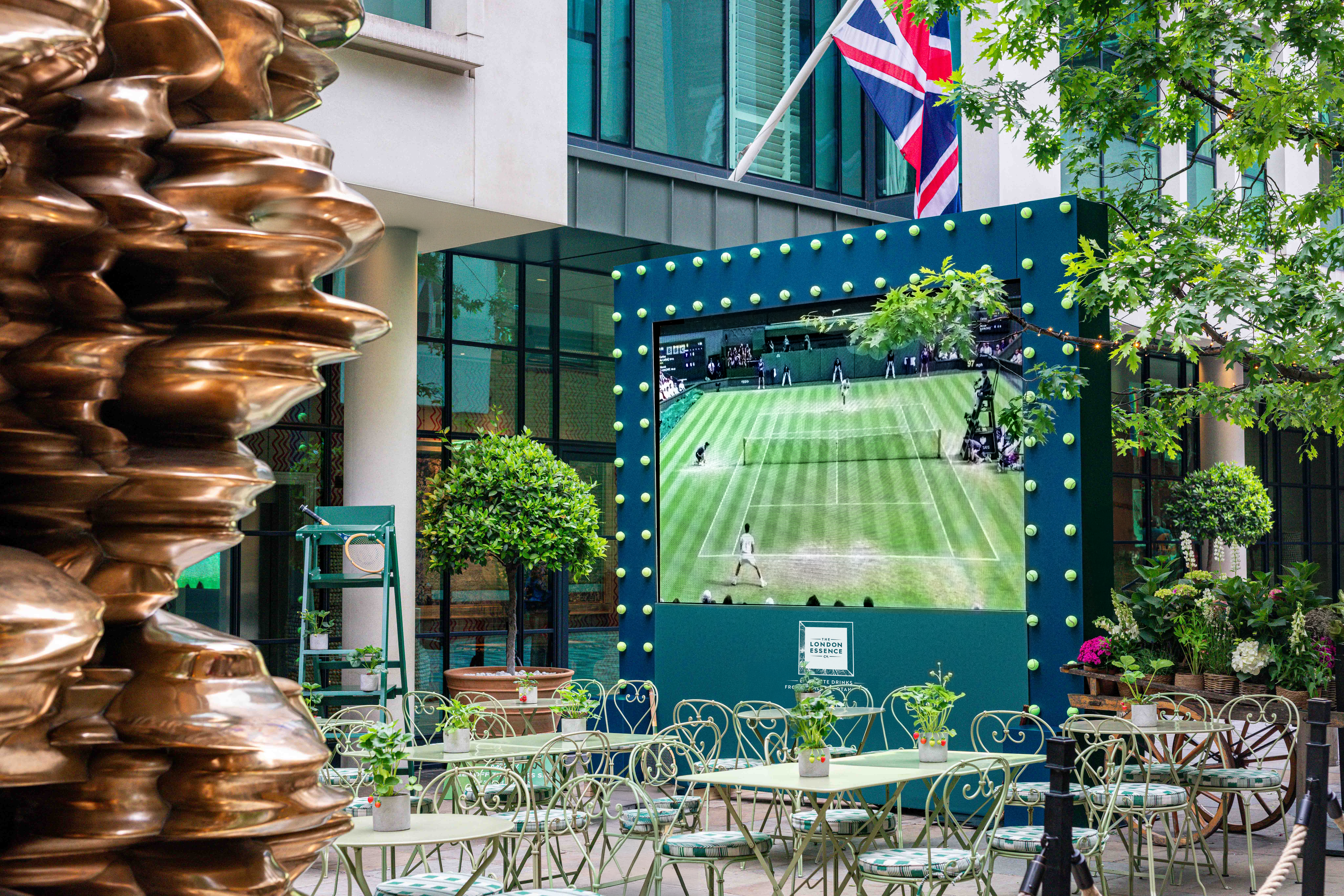An image of the big screen set up on the Ham Yard Hotel's courtyard displaying Wimbledon, with green tables and chairs in front of it