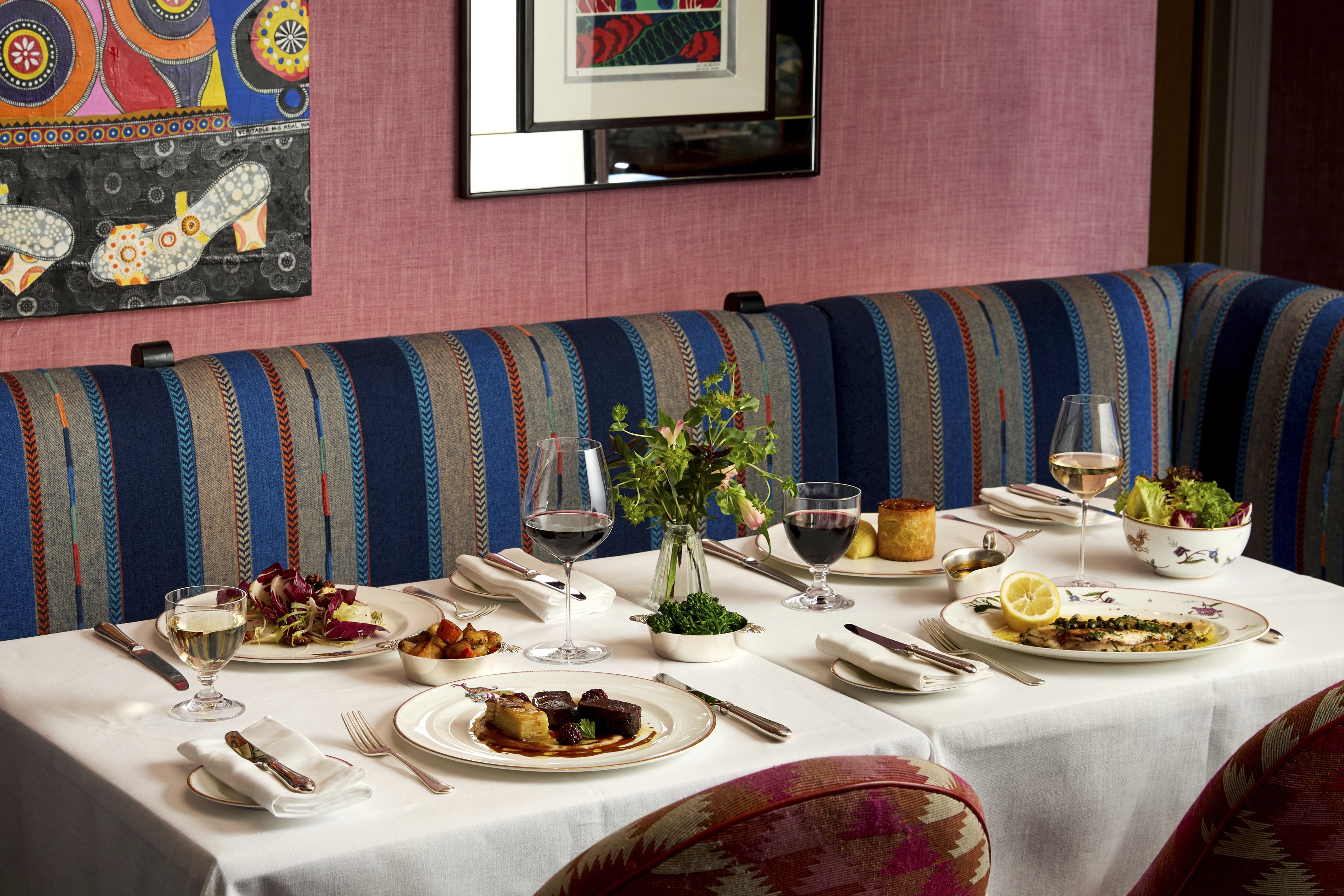 A table of four laid with main dishes accompanied by glasses of wine at Brumus. The table is positioned in front of banquette seating with art in partial view hanging on the wall