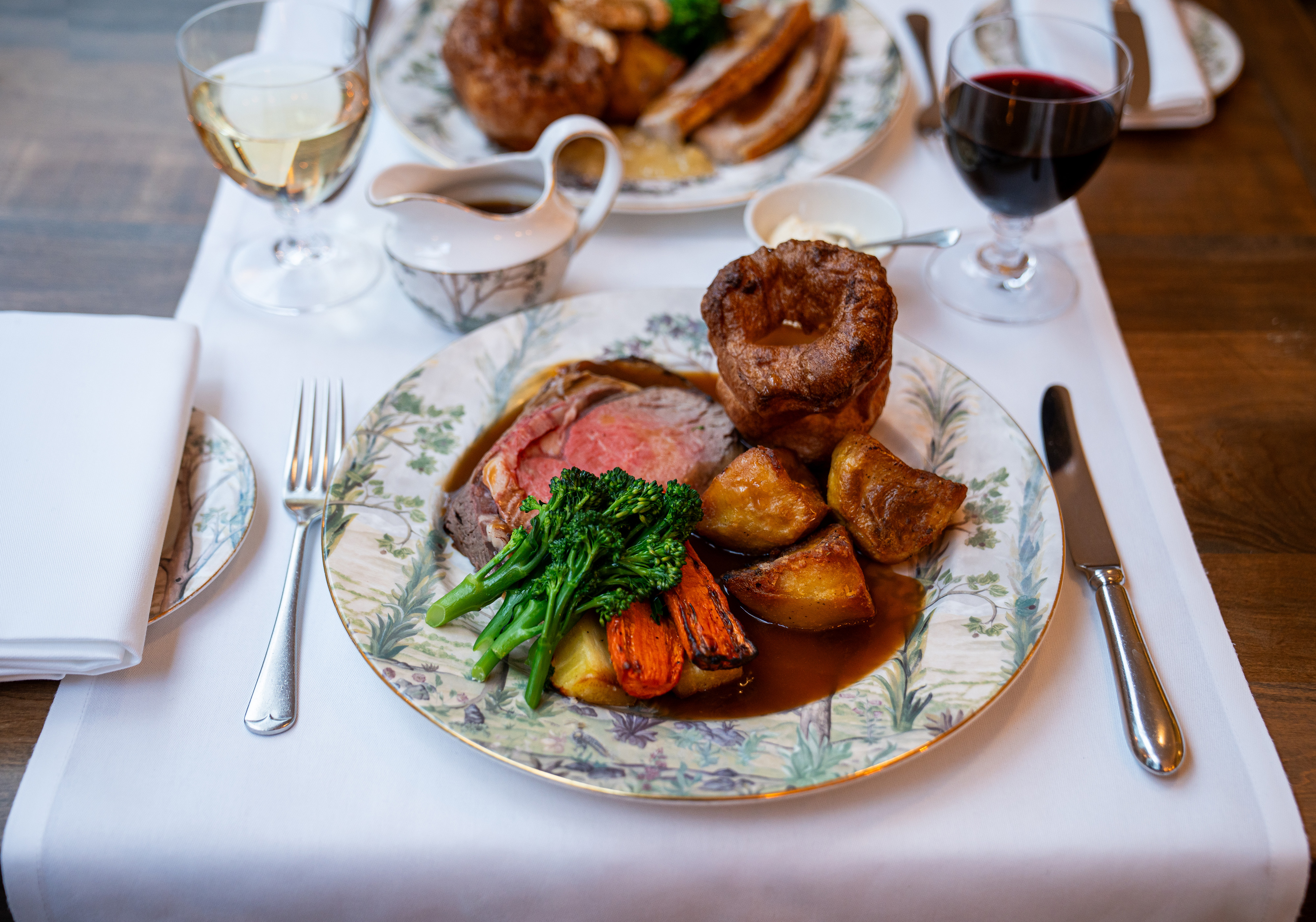 Sunday Roast at The Potting Shed. A plate of roast beef, with Yorkshire pudding, potatoes, vegetables and gravy with a plate of roast pork out of focus opposite. Both roasts are paired with glasses of wine