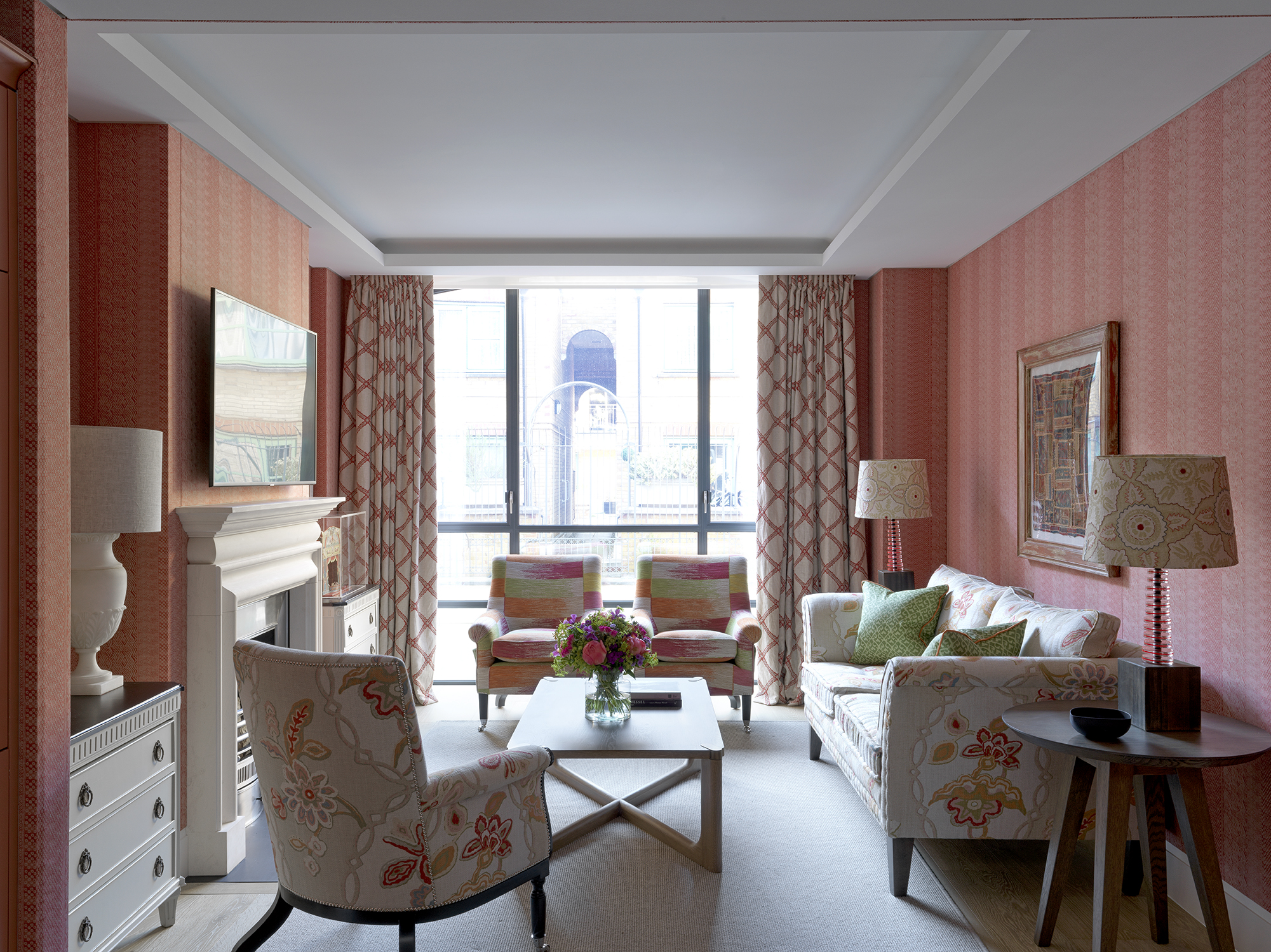 A wide shot of the living space are upholstered in a soft red fabric and a sofa, two armchairs and a coffee table frame a fireplace on the left hand side of the room. At the end of the room are floor-to-ceiling windows of the 9-11 Richmond Buildings.