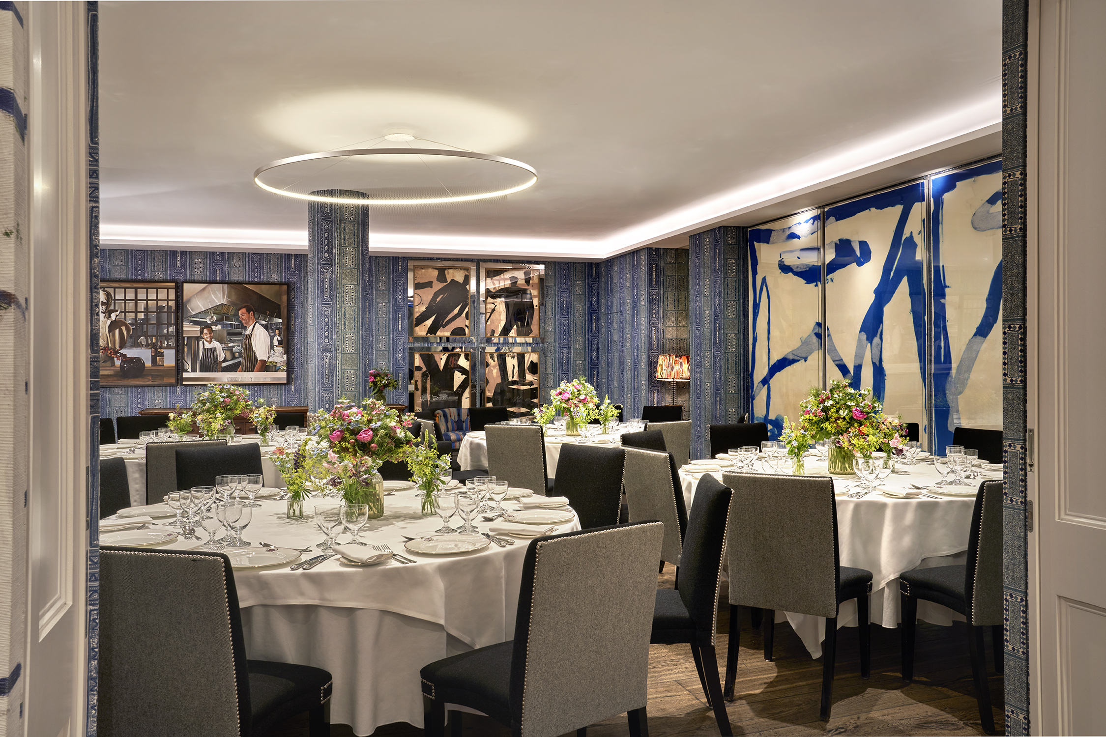 An image of multiple round dining table set up in the Indigo Room, there are white table cloths, floral bouquets in the centre and candles. each place around the table has a placemat, plate, cutlery, glassware and napkins ready for dining. private events room at The Soho Hotel.