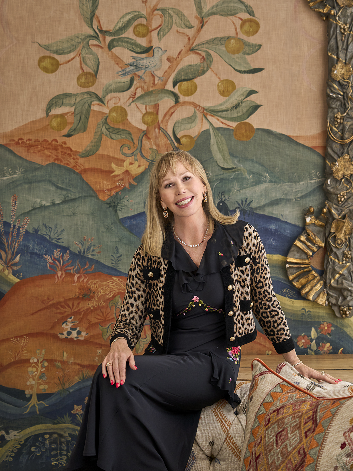 A portrait image of Kit Kemp in front of a wall covered in Kit Kemp x Andrew Martin Meadow Hillside, in the Drawing Room at The Soho Hotel