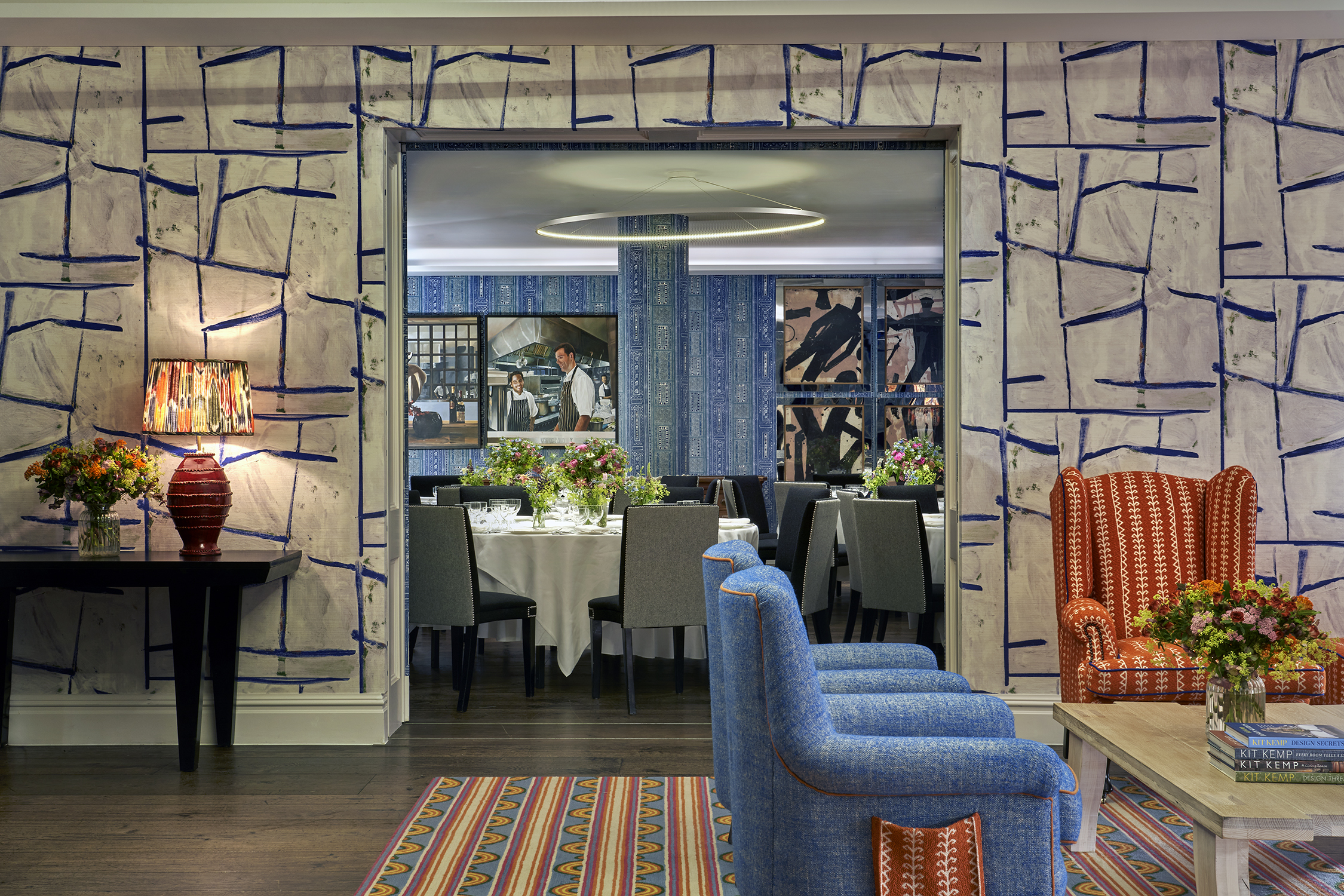 An image of all the Sandra Blow and Indigo private events rooms at The Soho Hotel. In the for ground of the image we see the back of two blue armchairs pointing towards the right, in the background we see through the open sliding doors into the Indigo Room which is set up with round tables with white tablecloths, placemats, plates, cutlery, glassware and flowers in the centre.