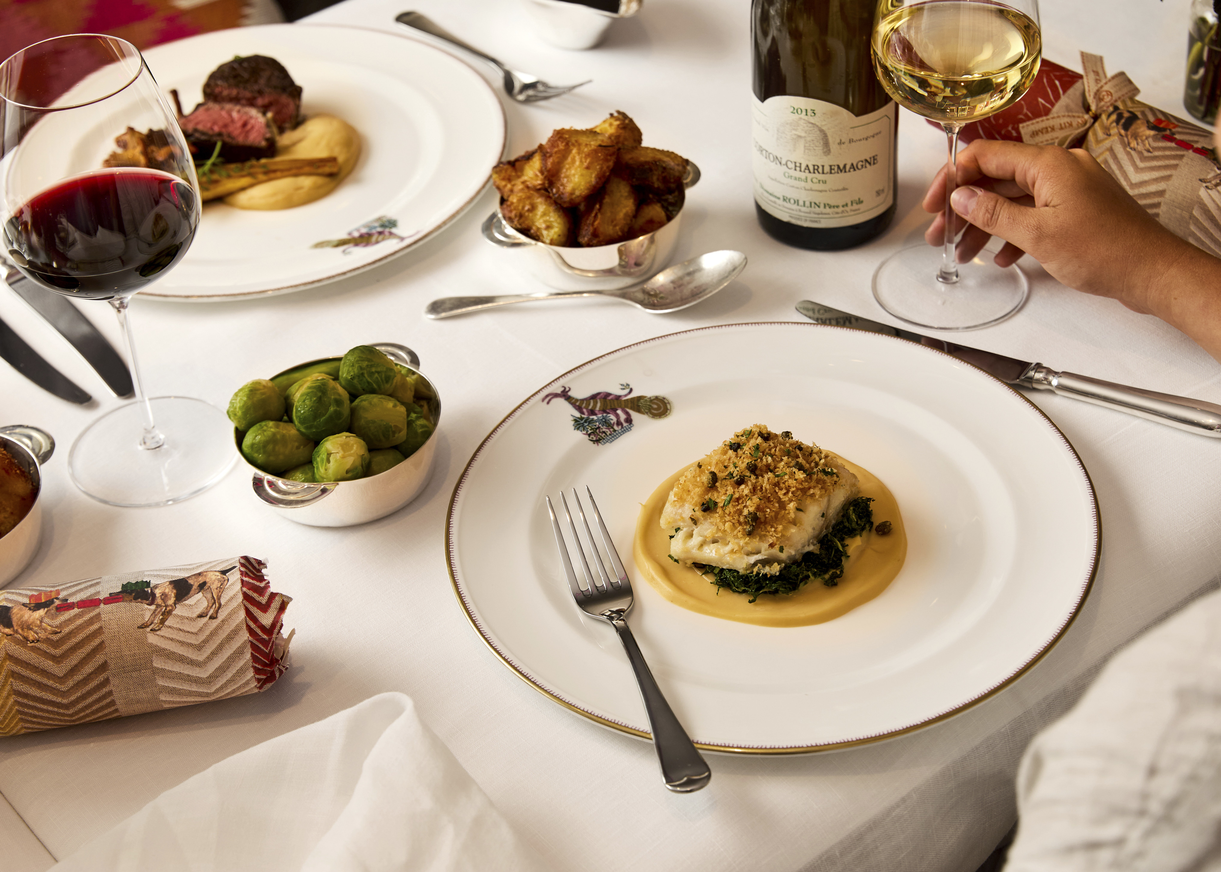 This image shows a festive Christmas meal in Brumus at Haymarket Hotel, with mythical creature crockery and dishes from the festive menu, including fish, steak, roast potatoes and Brussels sprouts, accompanied by red and white wine and Christmas crackers.