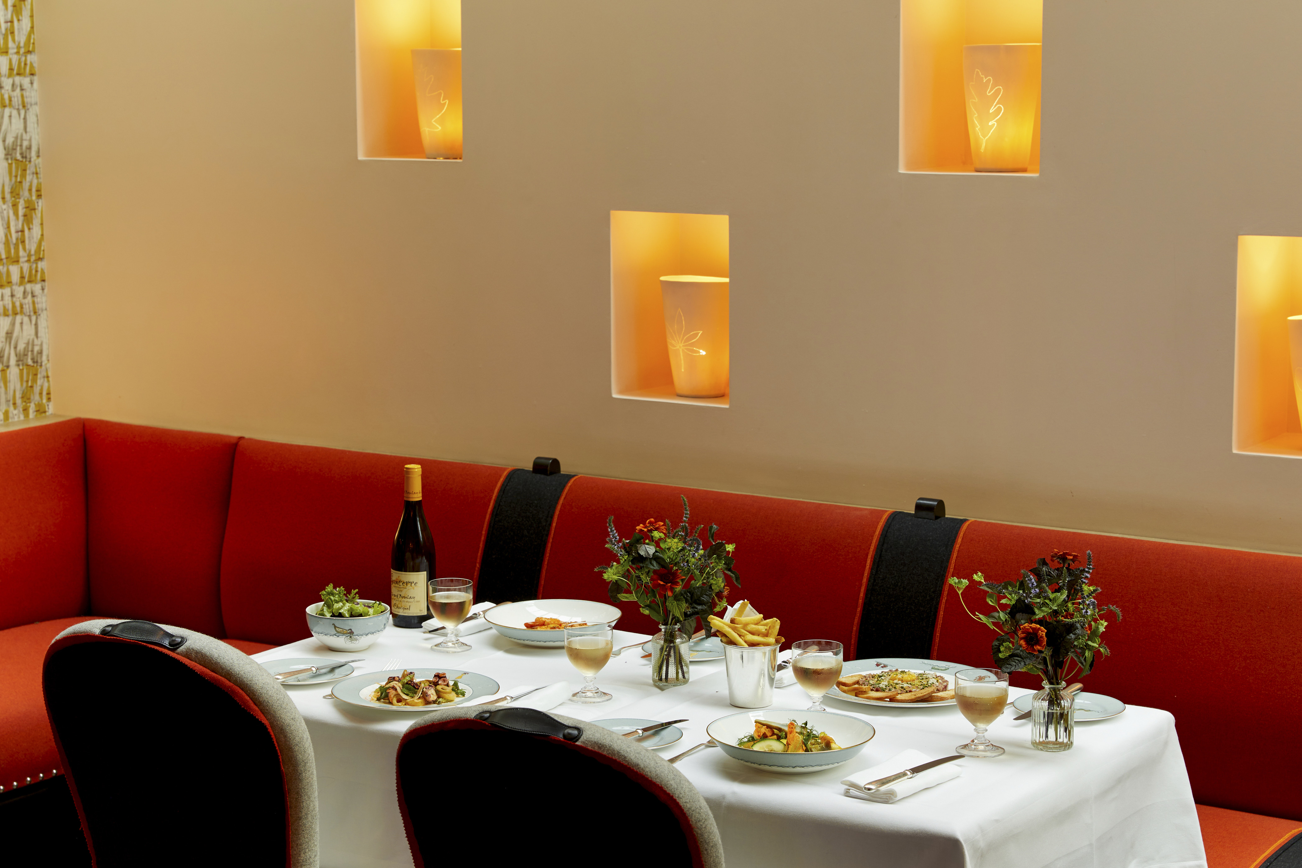 An image to promote the à la carte mains at Ham Yard Bar & Restaurant at Ham Yard Hotel. The image shows a white-linen table set with a selection of starters, including oysters, prawn cocktail, and vibrant salads. There are wine placed next to each dish, set against a backdrop of Martha Freuds ceramic installation.