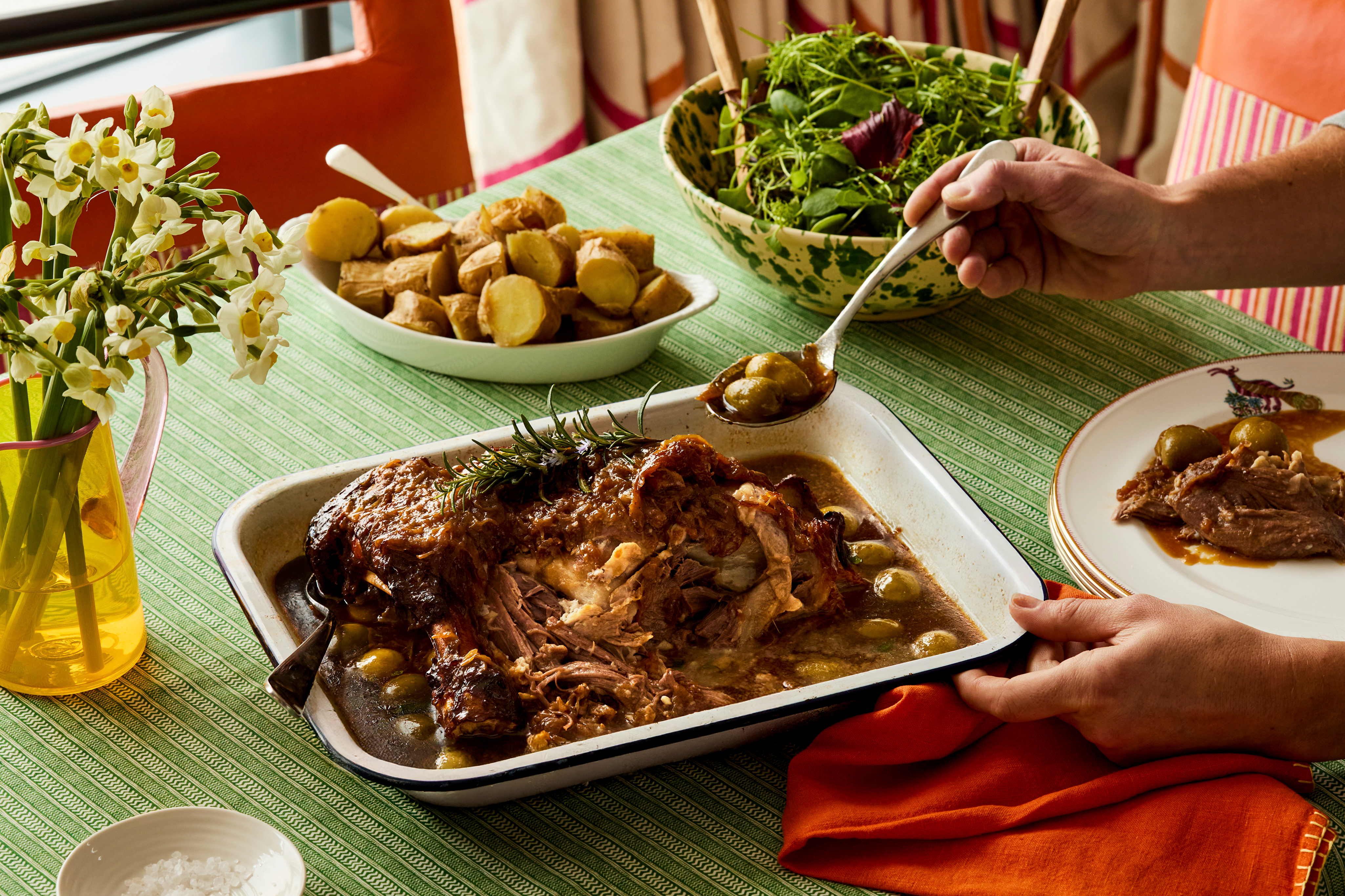 An oven dish filled with slow cooked lamb placed on a table with a hand taking a spoonful out to put on a nearby plate. The lamb is accompanied by potatoes and salad