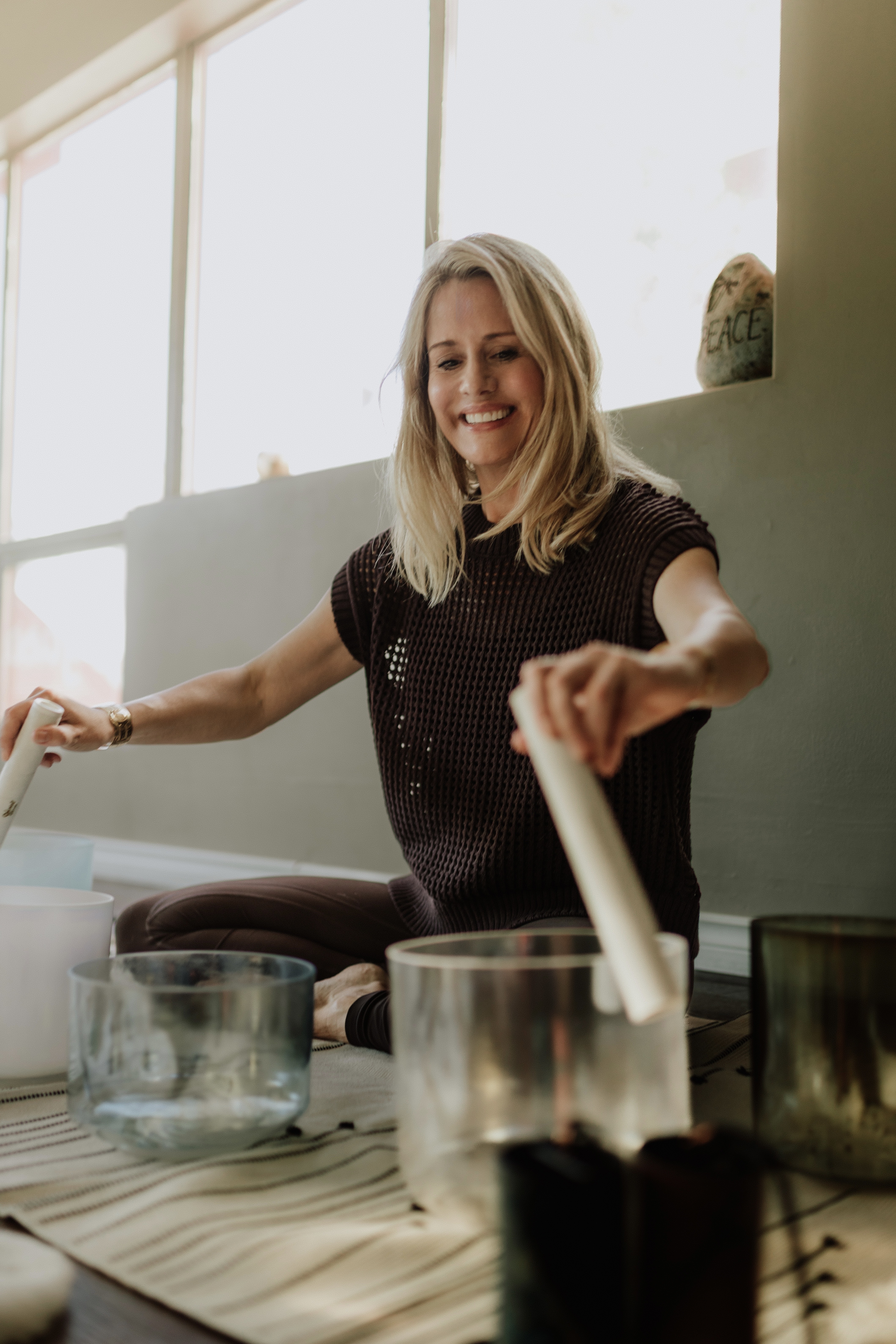 A close up shot of sound practitioner Nancy Trueman sitting on the floor playing crystal bowls with two large sticks