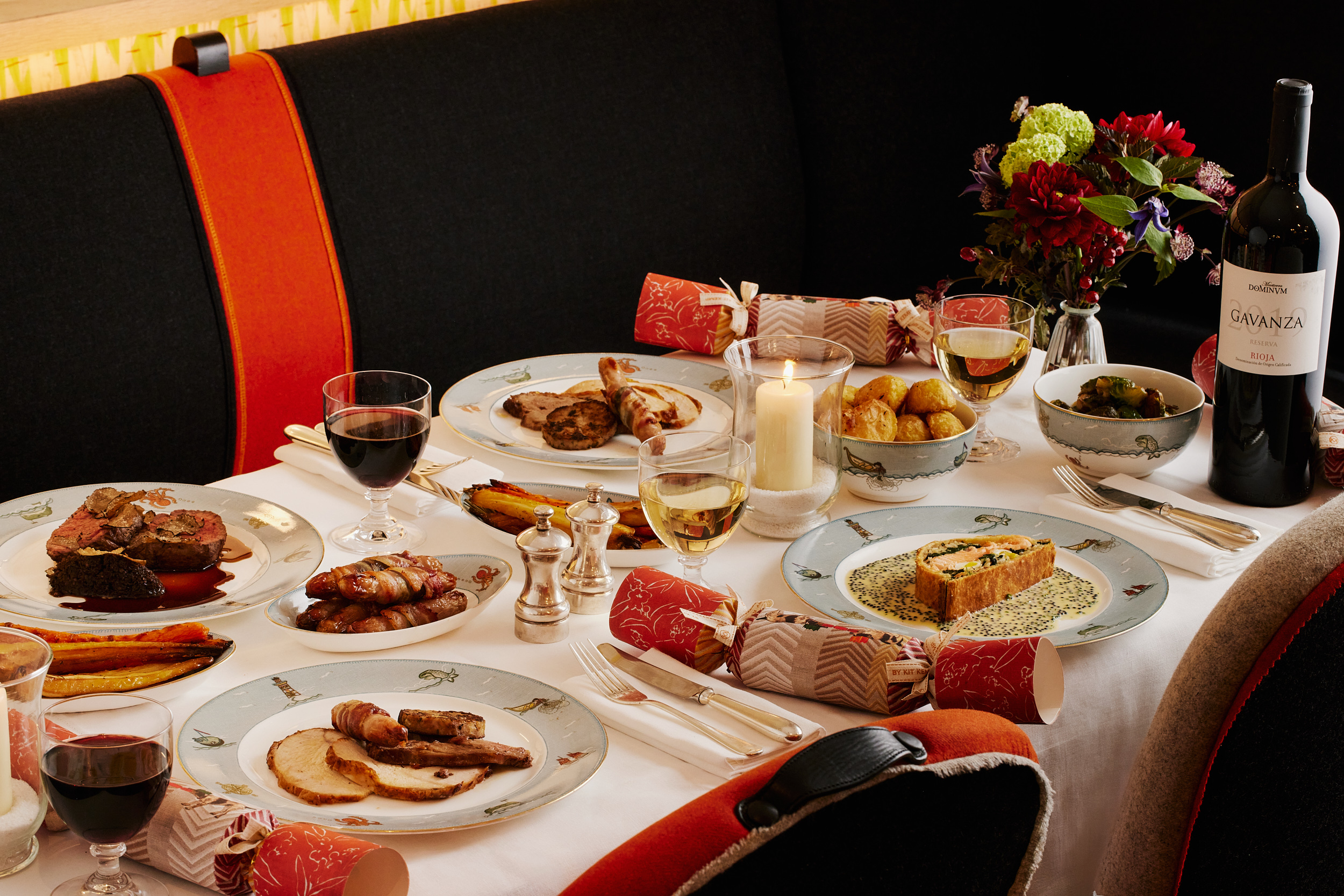 An image of the main dishes from the Christmas Day menu at Ham Yard Restaurant, Ham Yard Hotel. On the table, you can see all on Sailor Fairwell crockery. Also on the table you can see Roast Bronze turkey with all the trimmings, Beef Wellington, ceps and truffle duxelles, red wine sauce and the Salmon coulibiac, Exmoor caviar butter sauce. Also on the table there are side dishes of of roast vegetables, glasses of wine and champagne and festive flowers.
