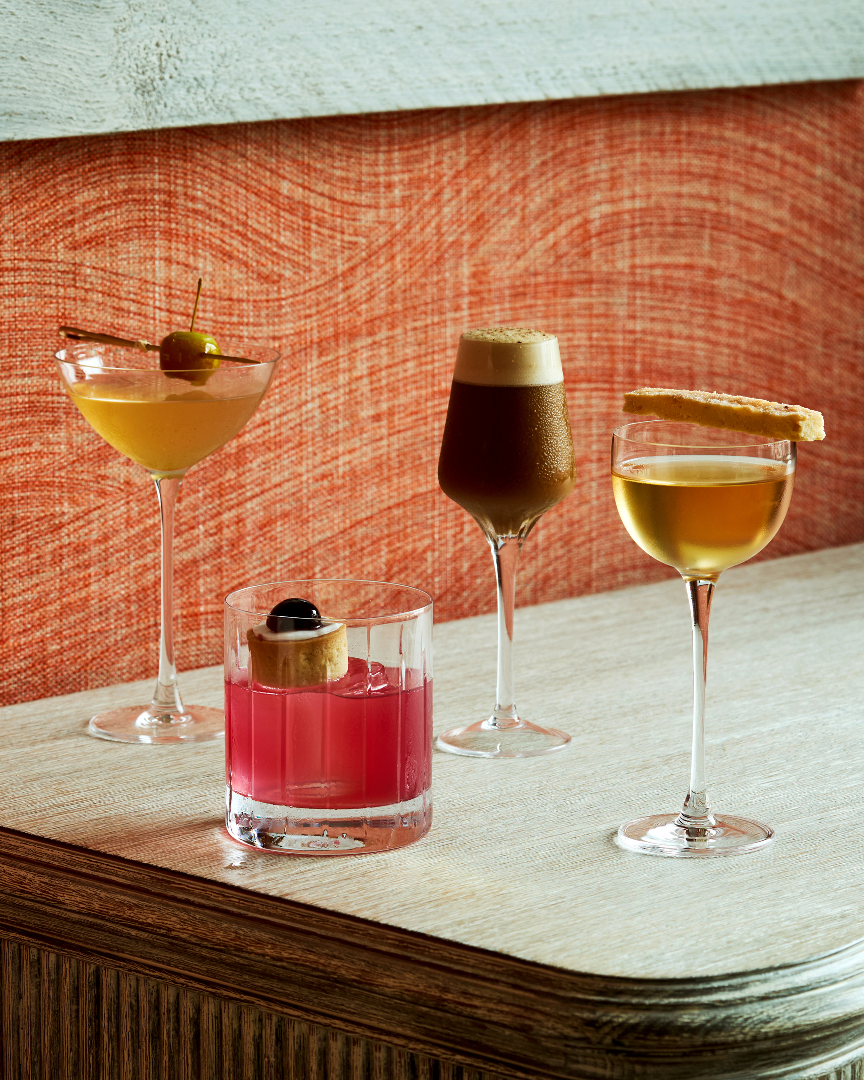 An image of 4 cocktails against a red fabric wall. 3 martini shaped glasses and one short whisky glass.