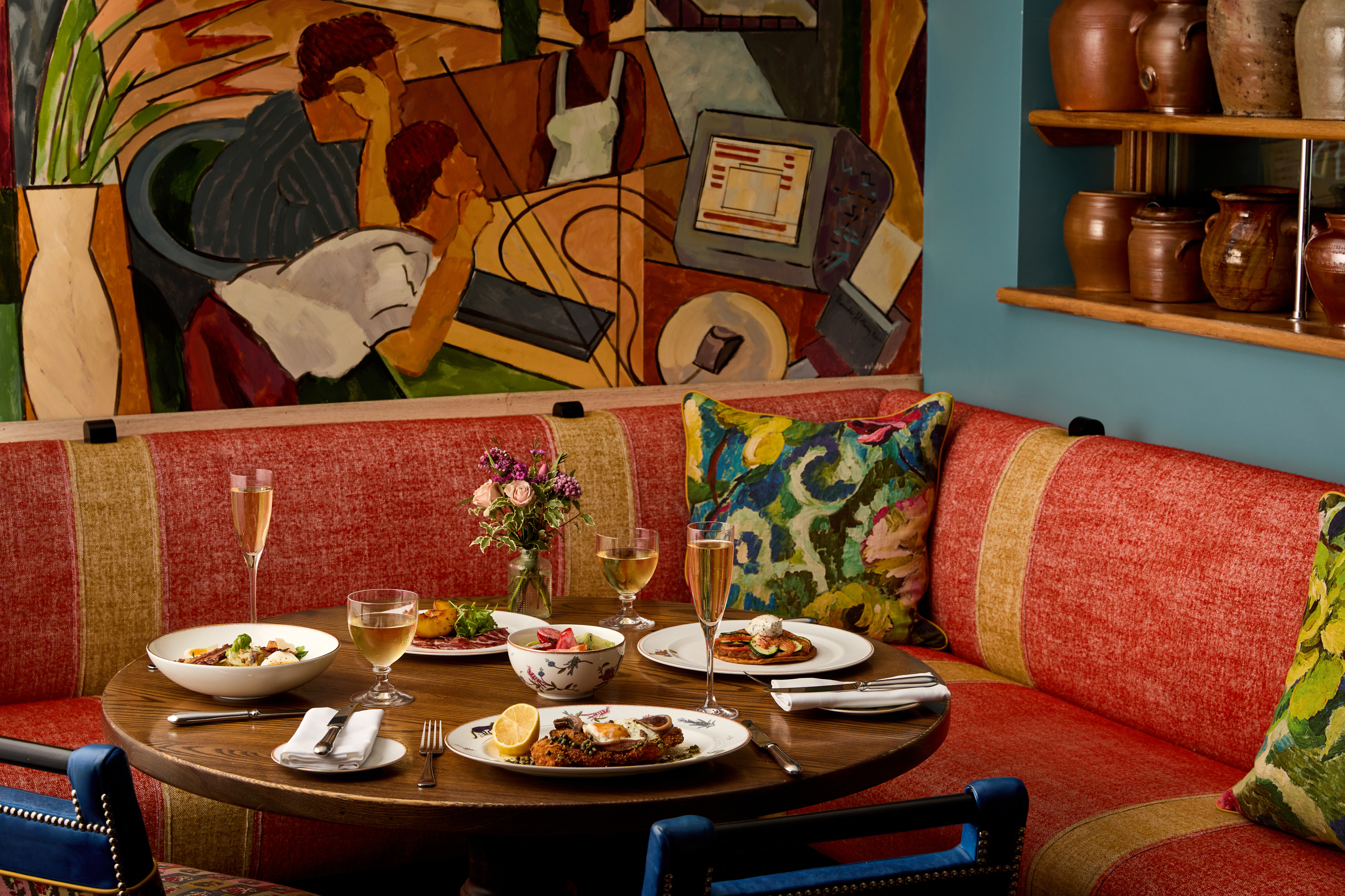 An image to promote Oscar Restaurant’s À La Carte menu at Charlotte Street Hotel. The image shows a round dark wood table with multiple dishes, examples of dishes include chicken schnitzel and trout dishes and with flutes of champagne and glasses of wine on the table.