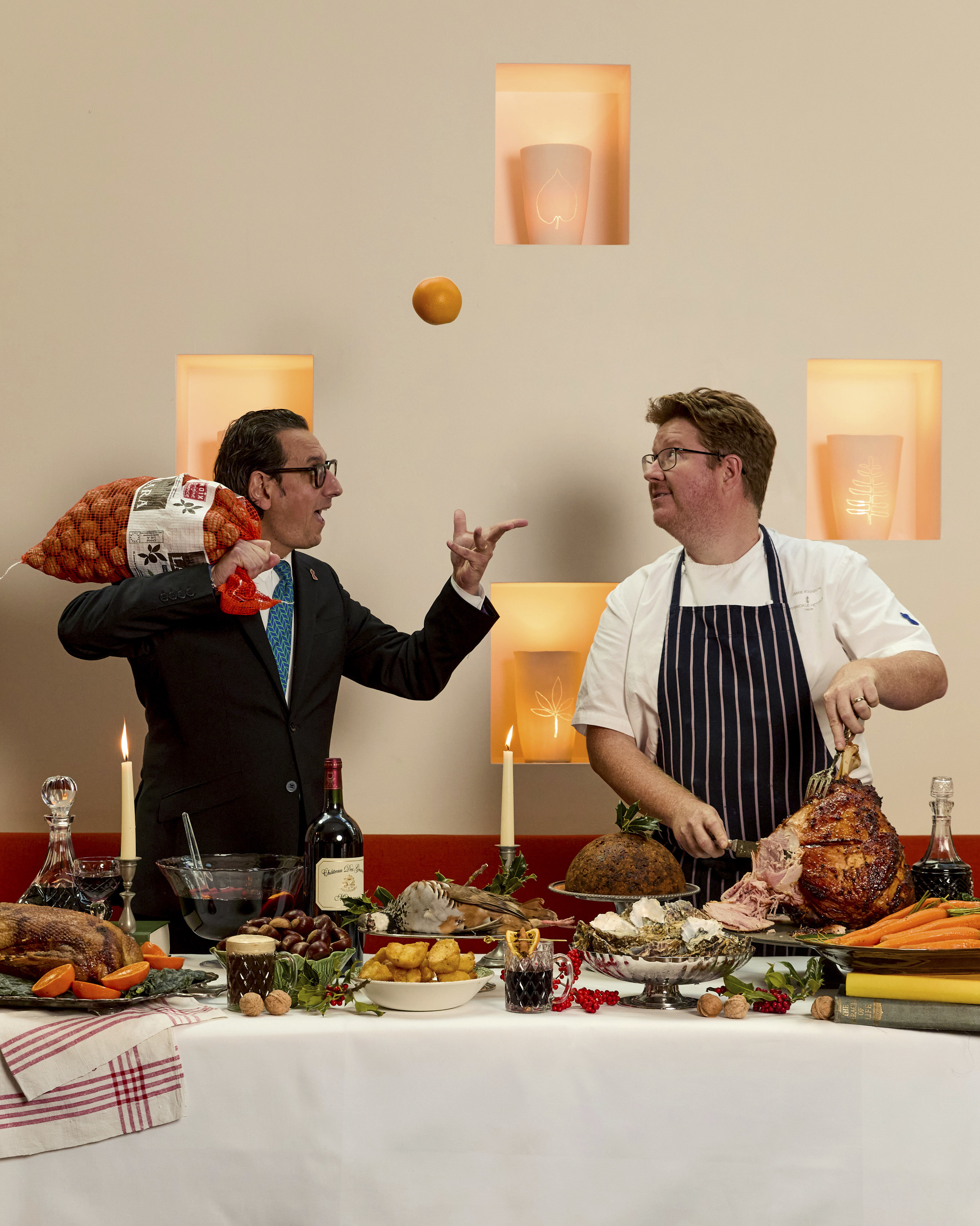 A portrait image of a large festive table spread filled with lots of different dishes including roast ham, roast duck, and a large Christmas pudding, bottle of wine and mulled wine, behind the table a sommelier throws and orange in the air as a chef carves the ham.