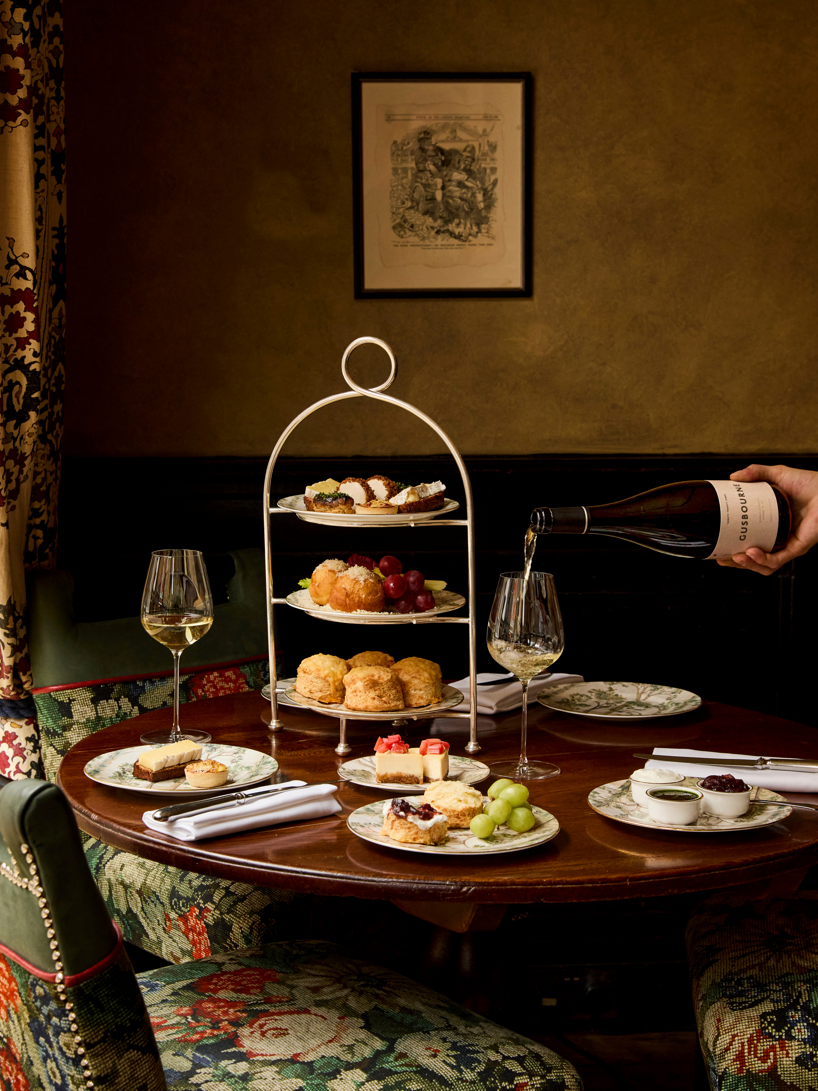 A portrait shot of the items in Afternoon Cheese at Brasserie Max at Covent Garden Hotel. On the table, there is a stand holding plates of scones and cheese-inspired pastries, and a bottle of white wine is being poured.