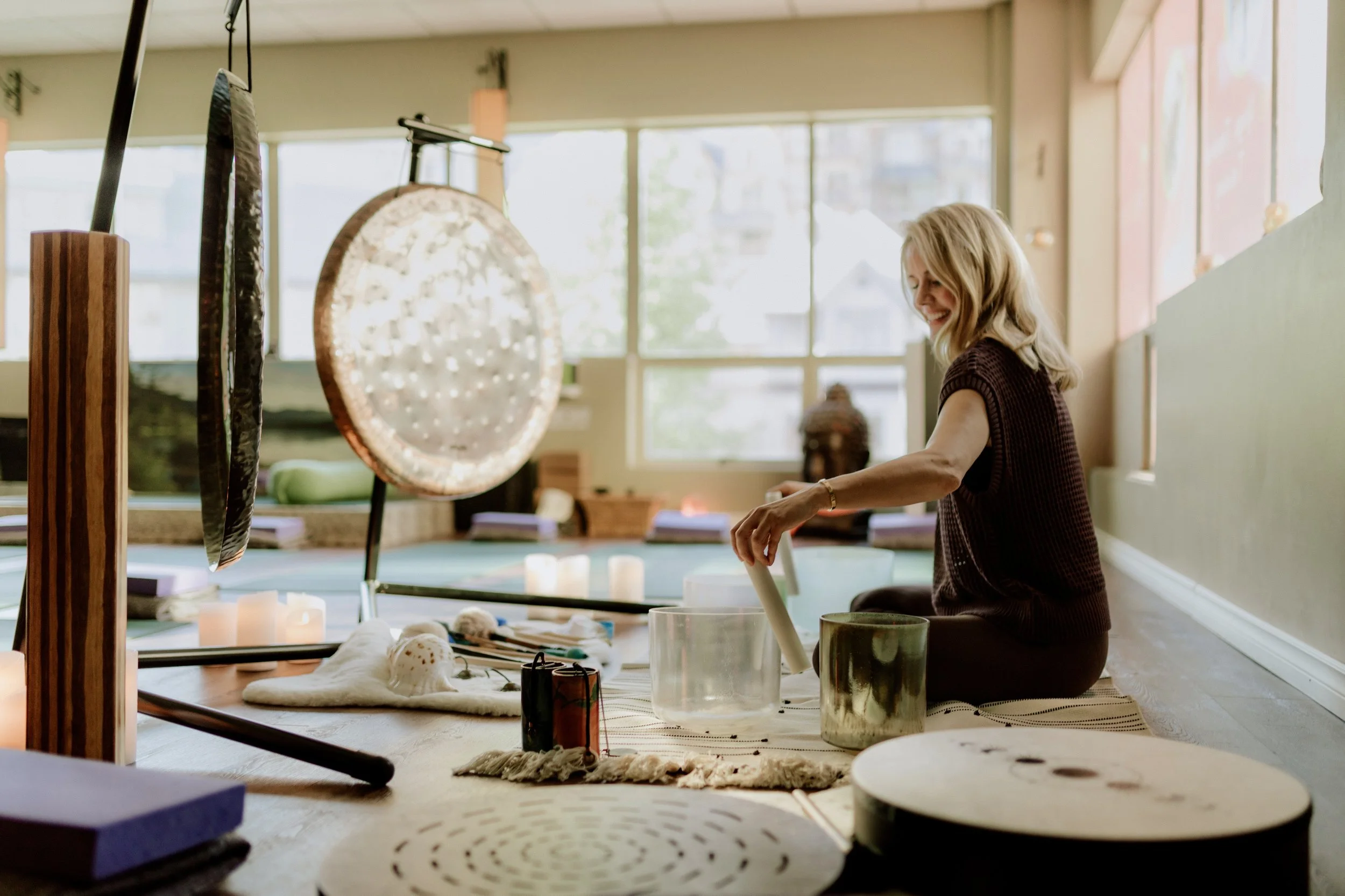 Sound practitioner Nancy Truman leading a sound bath class. She is sat on the floor playing with two glass sound bowls. In front of her are two large gongs
