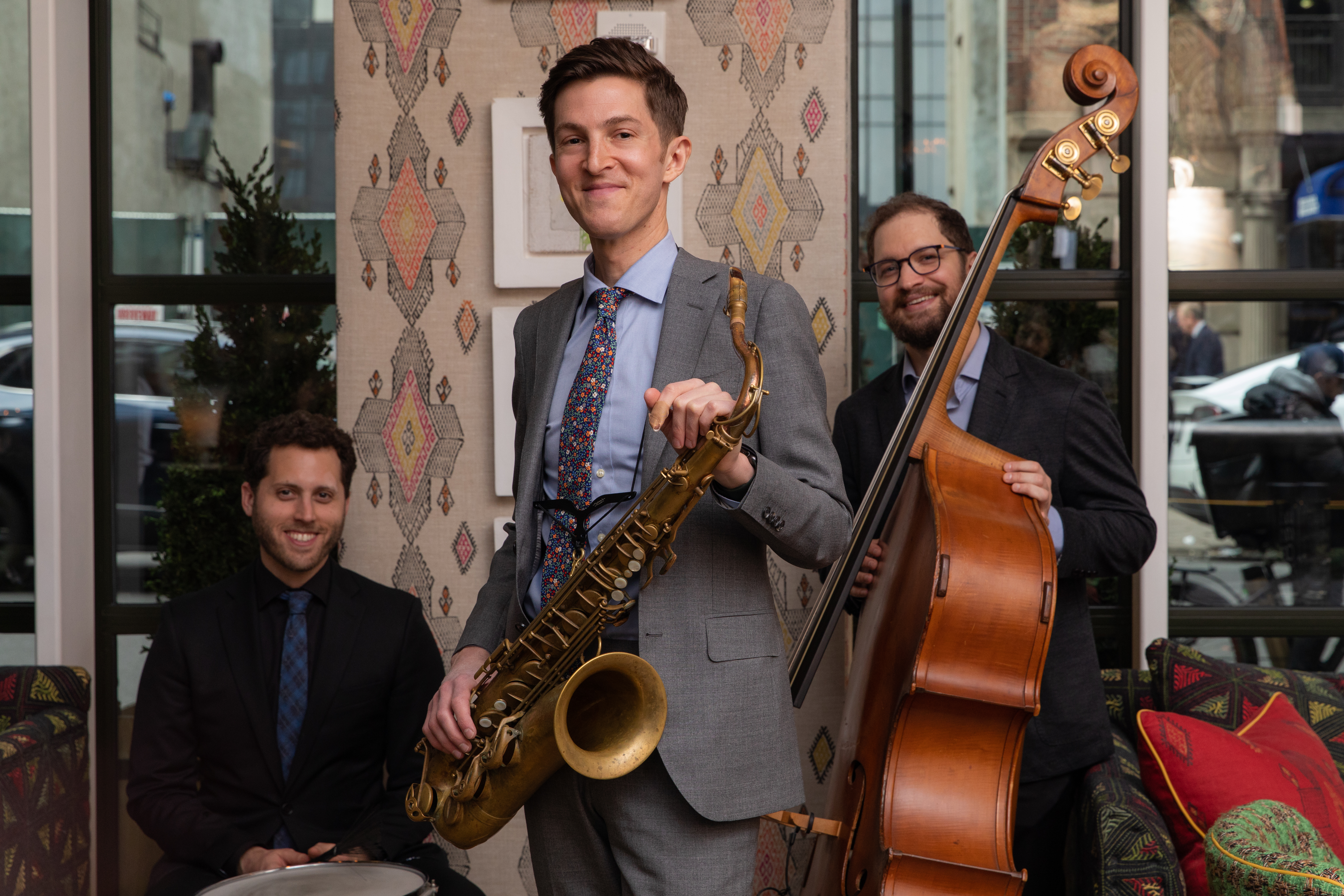 The Sam Taylor Jazz Trio. One man stands holding a saxophone in the middle, to the left there is man sat behind a drum and to the ridge, there is a man holding a bass