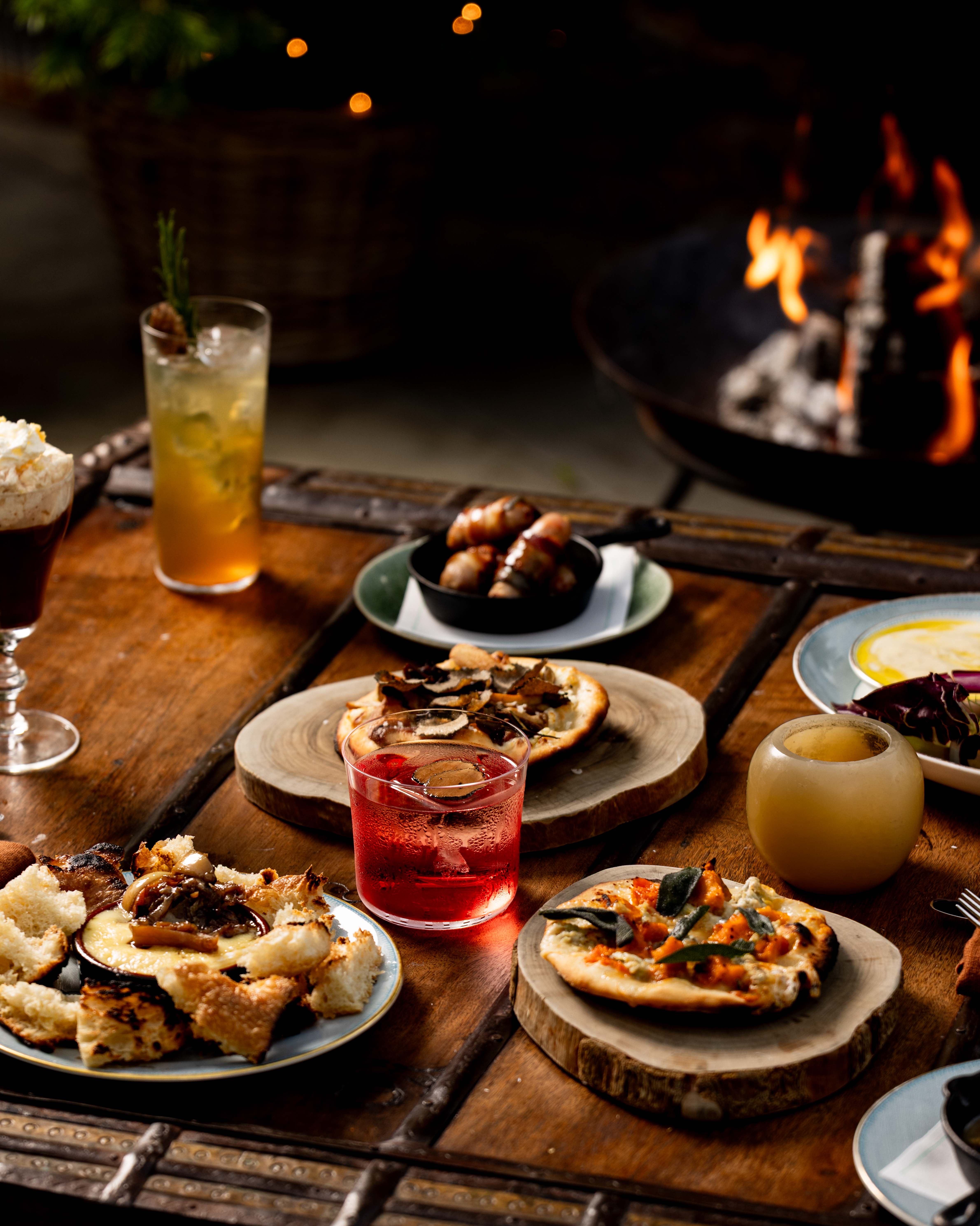 A portrait image a selection of fire-cooked dishes and cocktails from the Fire Garden menu at Ham Yard Hotel, they sit on a wooden coffee table