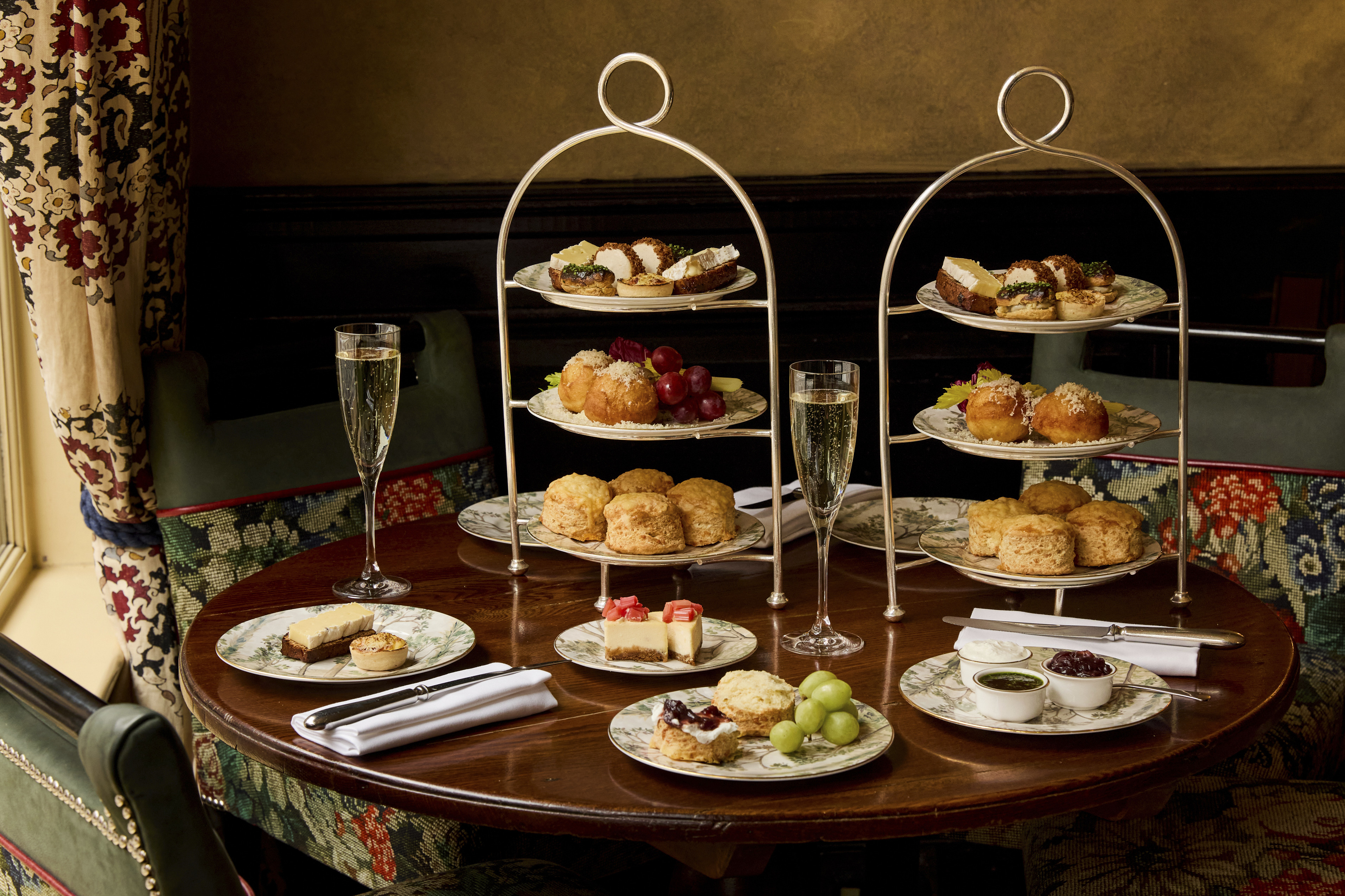 A wide shot of the items in Afternoon Cheese at Brasserie Max at Covent Garden Hotel. At the table, there are two stands holding plates of scones and cheese-inspired pastries, served alongside two glasses of Champagne.