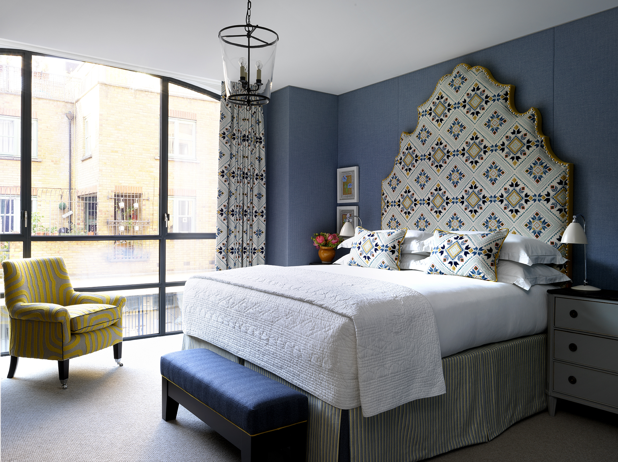 A wide shot of the view of the one bedrooms , A large statement headboard and bed dressed in crisp white sheets rest against walls upholstered in a dark navy fabric. There are floor to ceiling windows and a patterned yellow armchair in front of the windows at 9-11 Richmond Buildings.