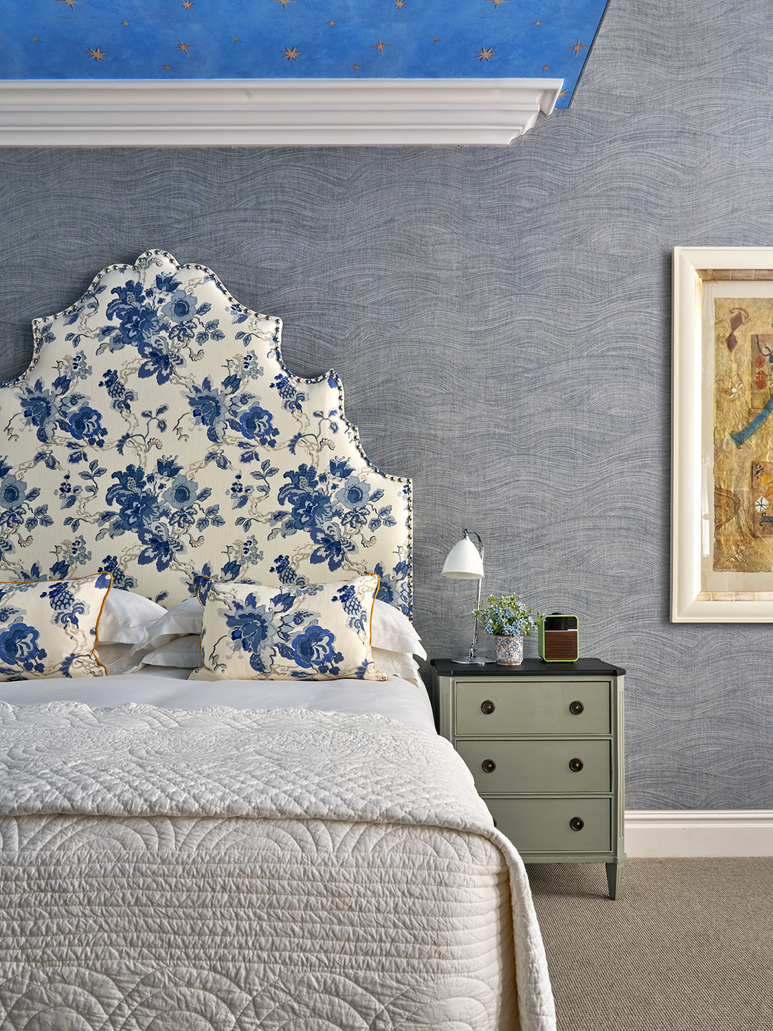 A portrait image focusing on the bed in The Knightsbridge Suite at Knightsbridge Hotel. In this image we see the bed with white sheets and pillows and decorative white and blue statement headboard with matching decorative pillows. To the right, we see a green bedside table with shelves.