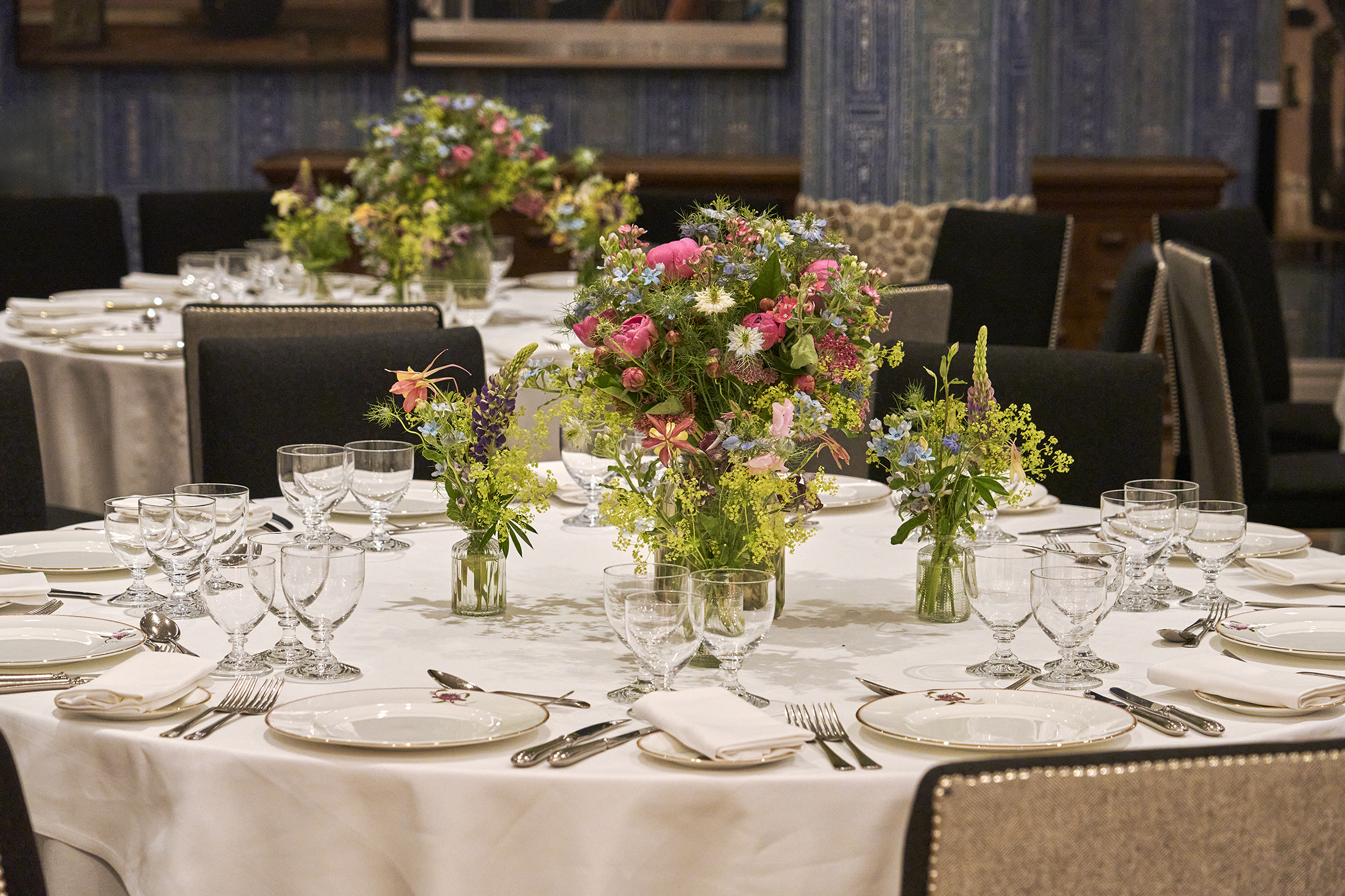 An close up image of the round dining table set up in the Indigo Room, there are white table cloths, floral bouquets in the centre and candles. each place around the table has a placemat, plate, cutlery, glassware and napkins ready for dining. private events room at The Soho Hotel.