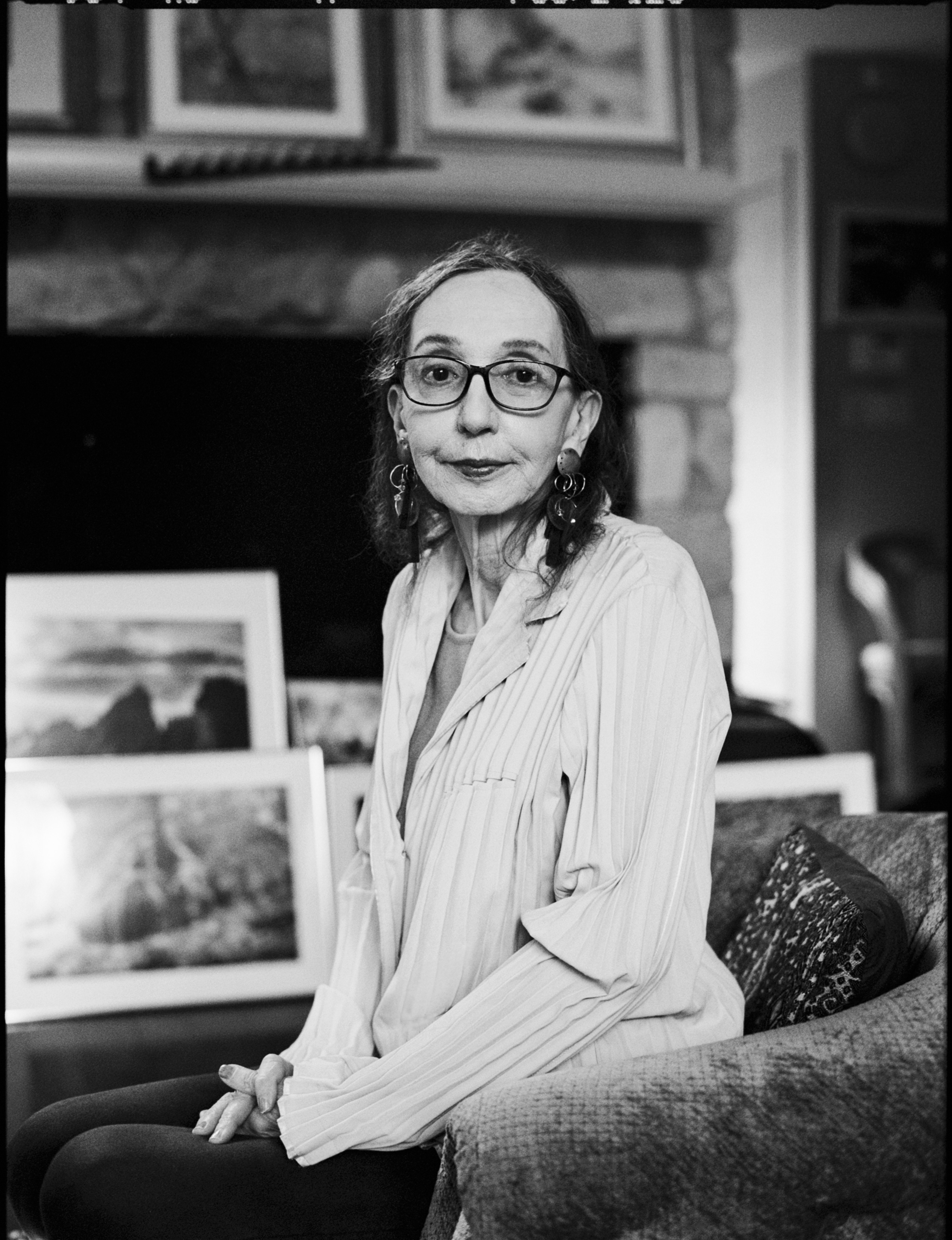A black and white headshot of author Joyce Carol Oates. She is sat on a sofa in a lounge, artwork can be seen out of focus behind her
