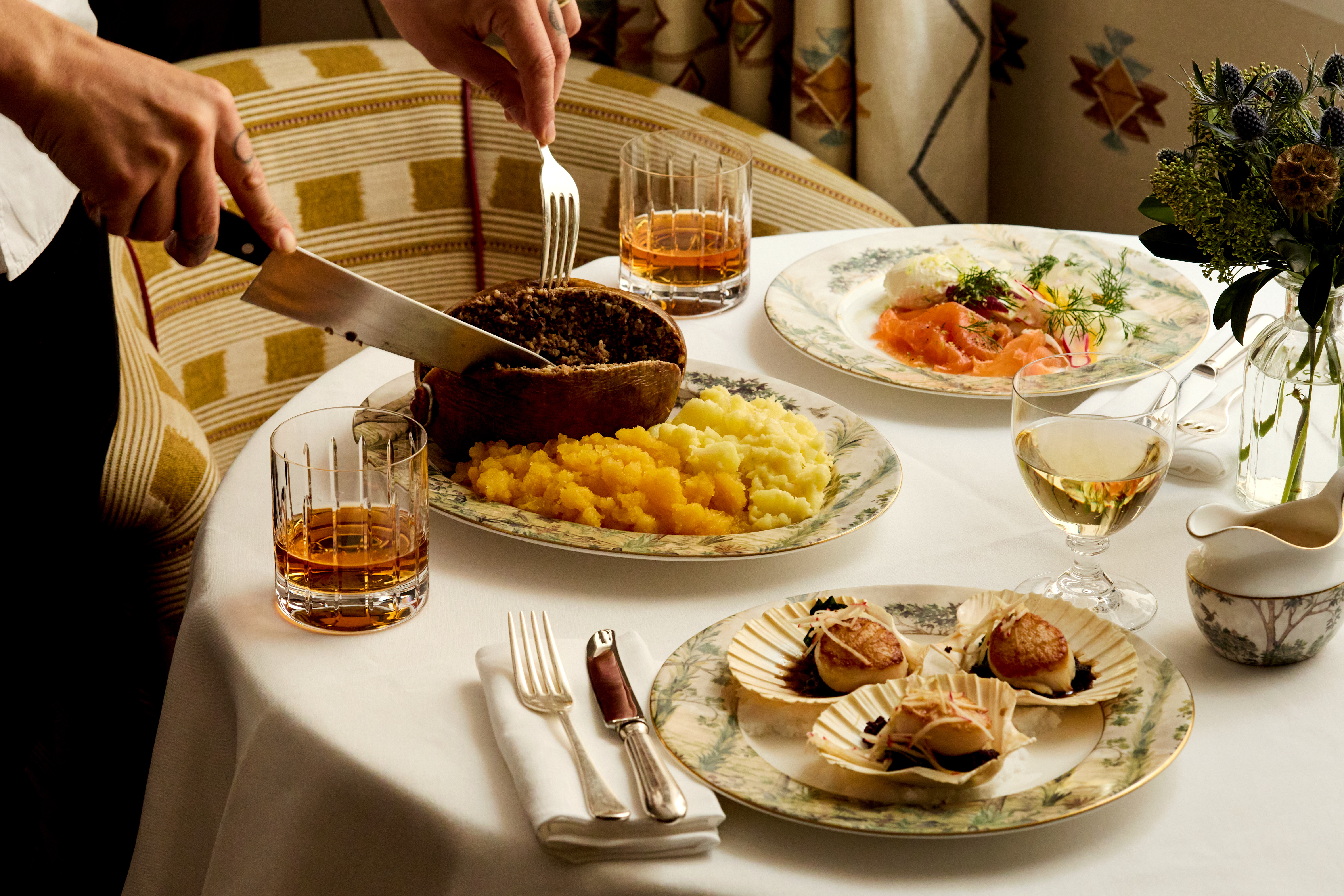 A table set for Burns Night dinner with Bruichladdich at Covent Garden Hotel. There is a plate of haggis being cut open, plates of scallops and smoked salmon and glasses of whisky.