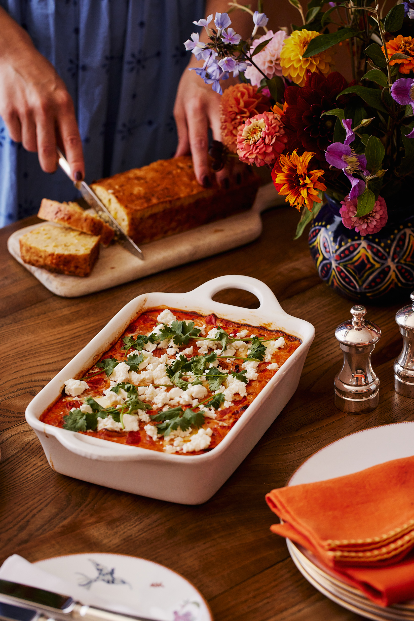 This images a portrait image that shows the final product of the recipe of Baked eggs and cornbread, here you can see a white baking dish with baked eggs in a totamto sauce topped with feta and corriander. in the bakground you can see a lady in a blue dress cutting into cornbread on a wooden board. in the foreground there is a stack of mythical creatures side plates and an orange napkin from Kit Kemp shop.