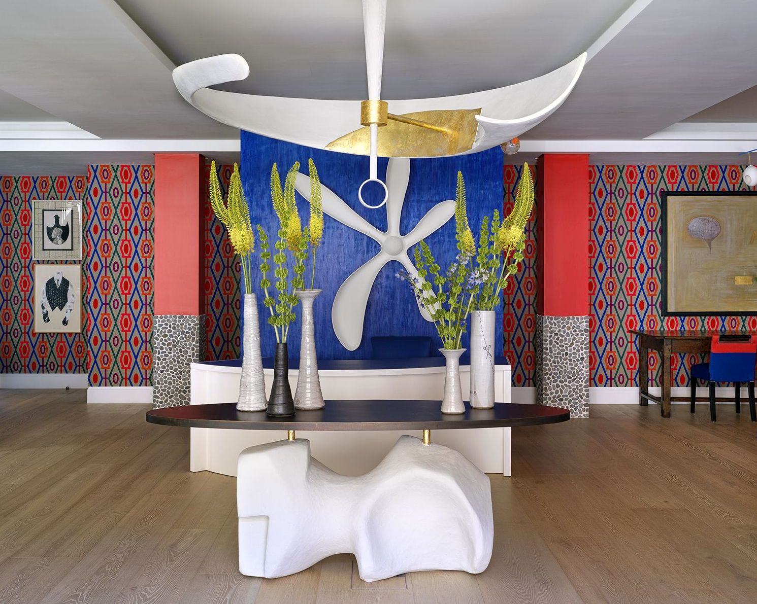 A wide shot of the lobby reception area featuring a central table and a large sculptural light hanging above. In the background is a reception desk with a large sculptural flower hung on the blue wall behind. The lobby area is bright and colourful with patterned fabric adorning the walls and statement pieces of artwork at 9-11 Richmond Buildings