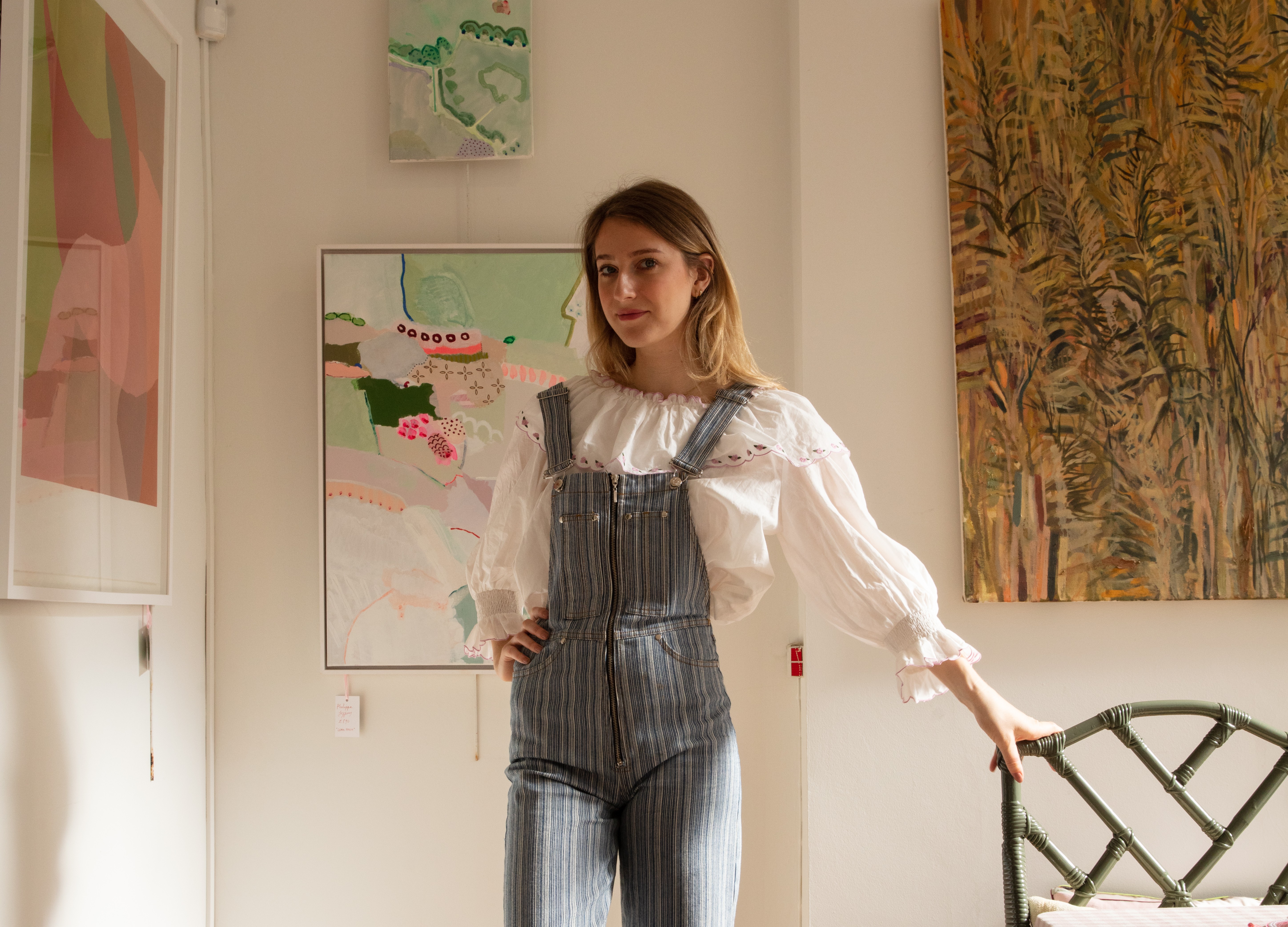 Artist and curator Domenica Marland dressed in dungarees, leaning on a small chair. Her framed paintings hang on white walls behind her