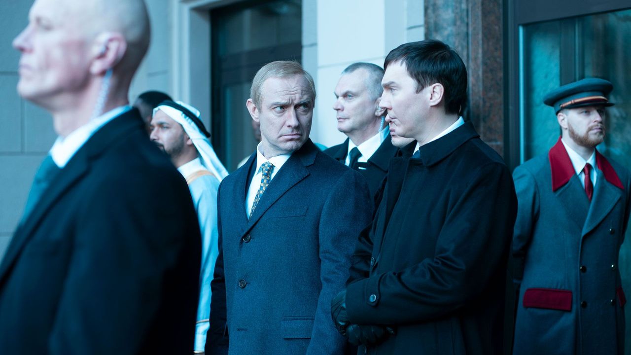 A still from the film "The Wizard of the Kremlin": A group of men are stood outside a building and one man wearing a navy blazer looks over at the man next to him