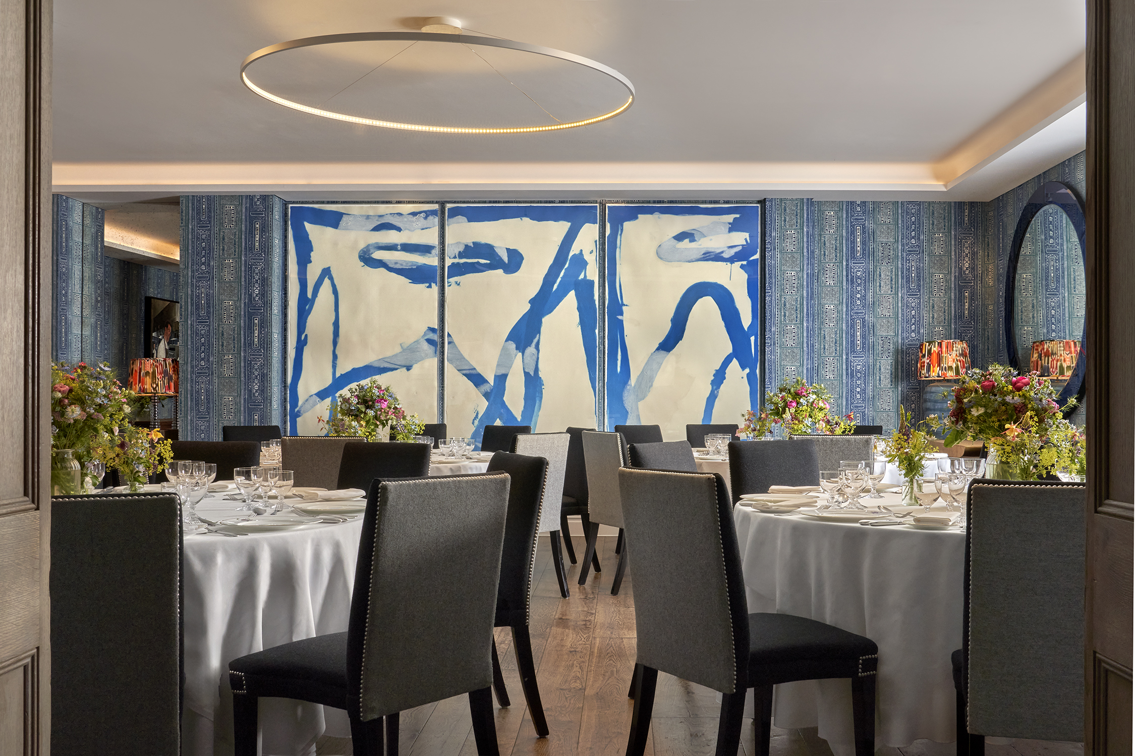An image of multiple round dining table set up in the Indigo Room, there are white table cloths, floral bouquets in the centre and candles. each place around the table has a placemat, plate, cutlery, glassware and napkins ready for dining. private events room at The Soho Hotel.