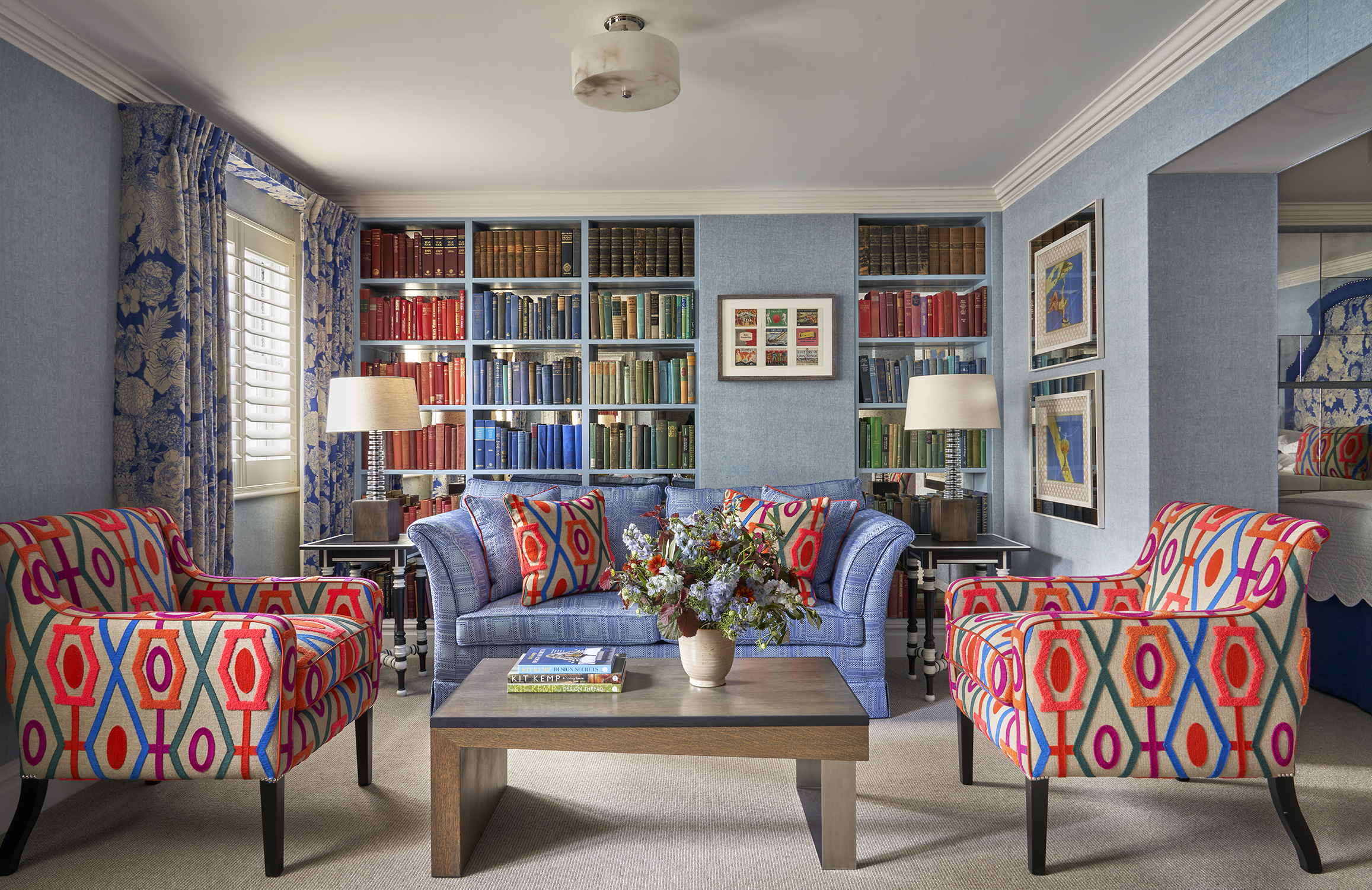 Seating area shot of room 15, the Marylebone Room at Dorset Square Hotel. The image shows a blue sofa in the centre in front of a colourful bookshelf wall full of books. In front of the sofa, there is a coffee table with patterned armchairs on either side.
