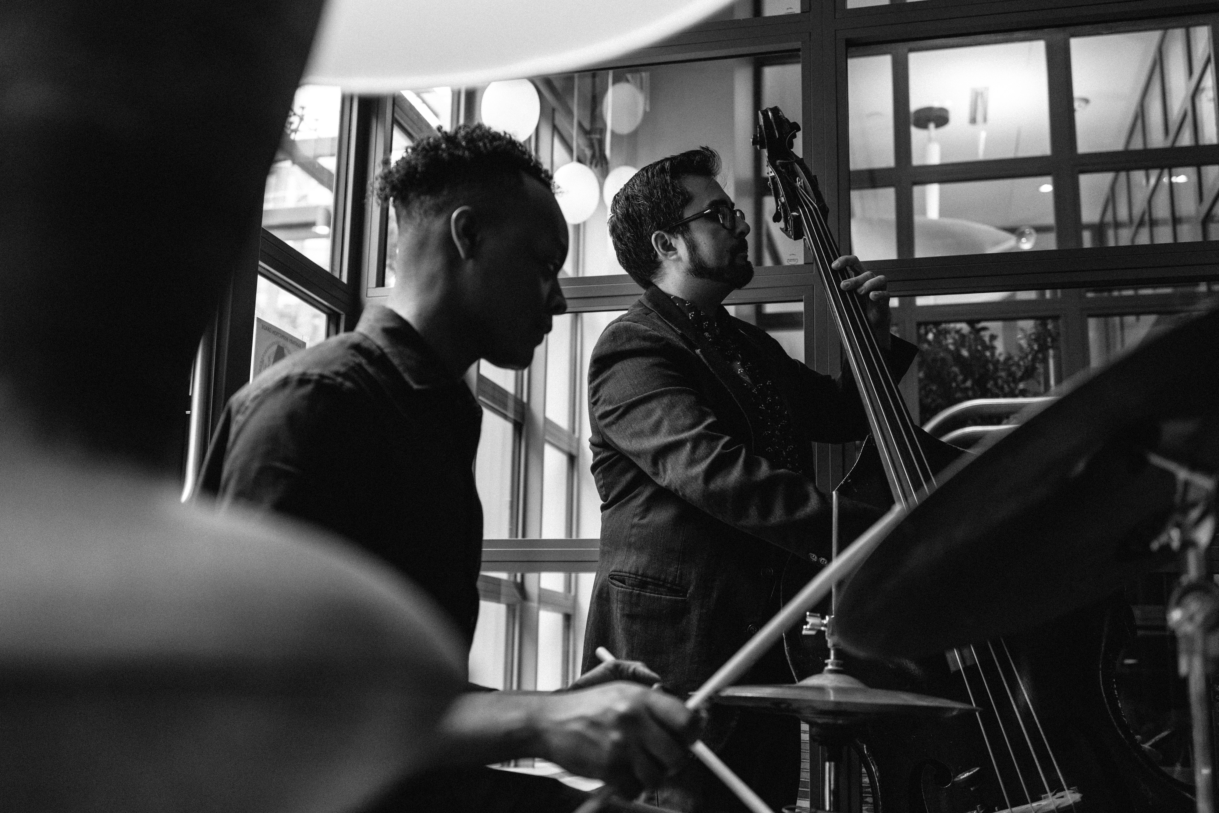 A black and white landscape image of a band playing jazz music, in a centre stands a guy with a upright bass and on the left a man playing the drums in the Warren Street Bar & Restaurant at Warren Street Hotel.