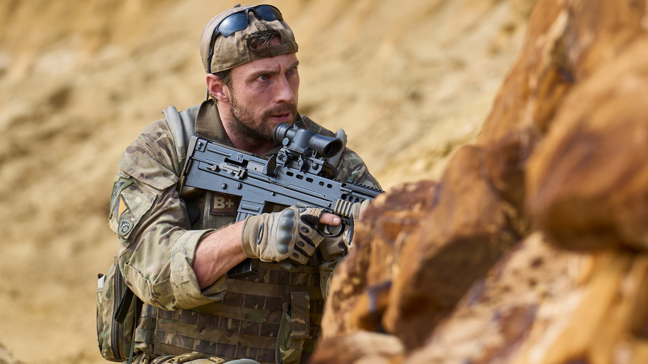 A still from the film 'Fuze'. A man dressed in camouflage army uniform crouches behind rocks in a desert setting, holding a large gun