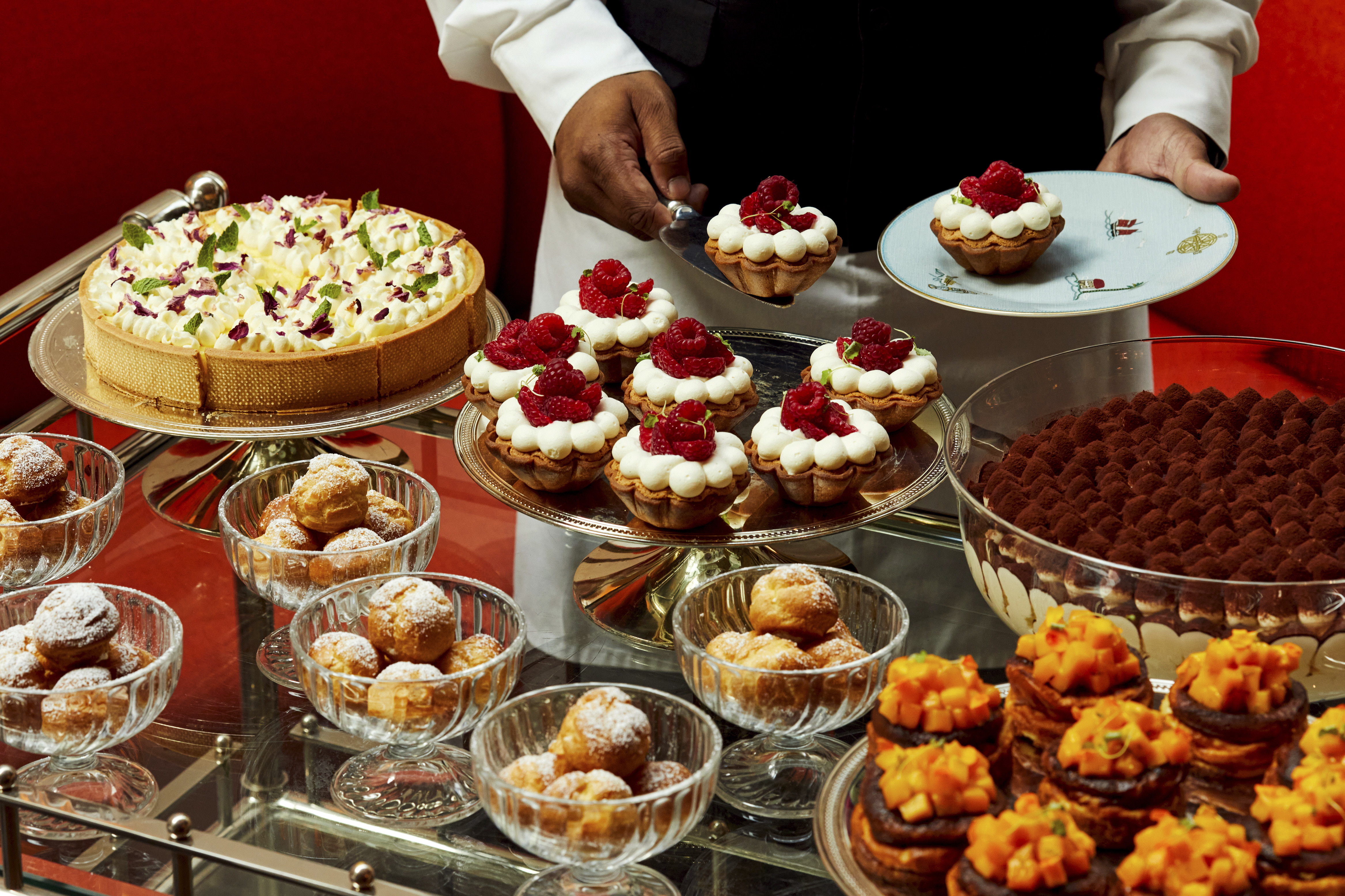 An image to promote the Long Lunch at Ham Yard Bar & restaurant at Ham Yard Hotel, The image shows a luxurious dessert trolley laden with an assortment of intricately presented pastries and cakes. A server in formal attire is plating a raspberry tart.