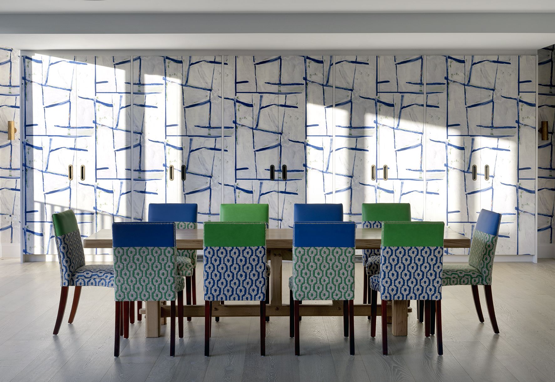 A view of the entertaining area on the top floor featuring a large wooden dining table and 10 chairs upholstered in colourful patterned fabrics. Behind the table are floor-to-ceiling cupboards covered in a graphic, geometric print at 9-11 Richmond Buildings