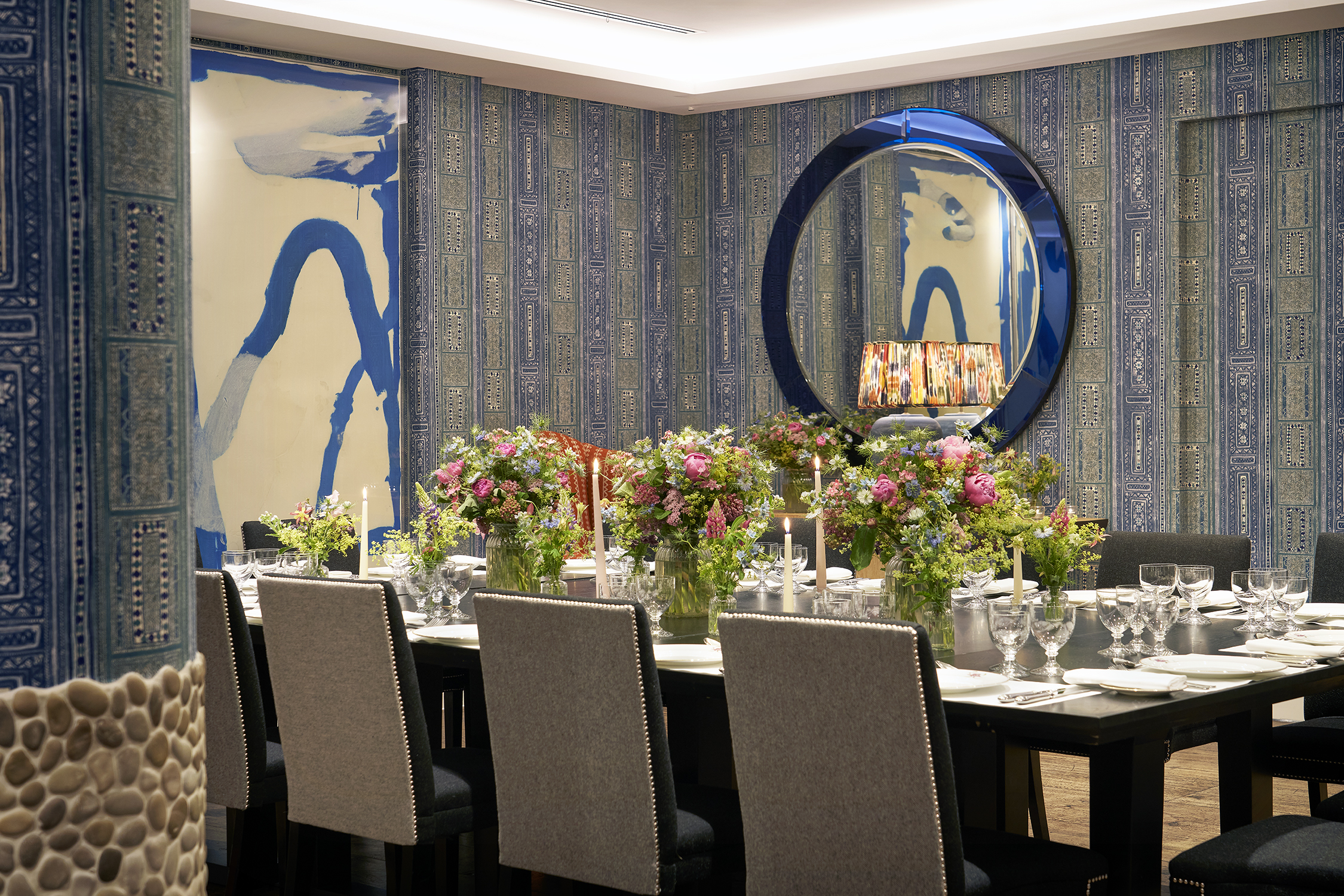 An image of the long dining table set up in the Indigo Room, there are floral bouquets in the centre and candles. each place around the table has a placemat, plate, cutlery, glassware and napkins ready for dining. private events room at The Soho Hotel.