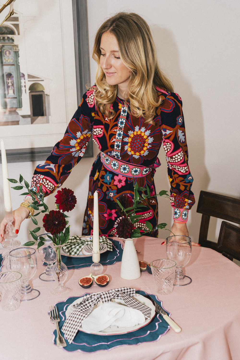 Artist and curator Domenica Marland dressed in a colourful patterned dress, creating a table scape. She is placing a candlestick on a dining table dressed with a pink tablecloth, plates, glasses, tablemats and small bud vases