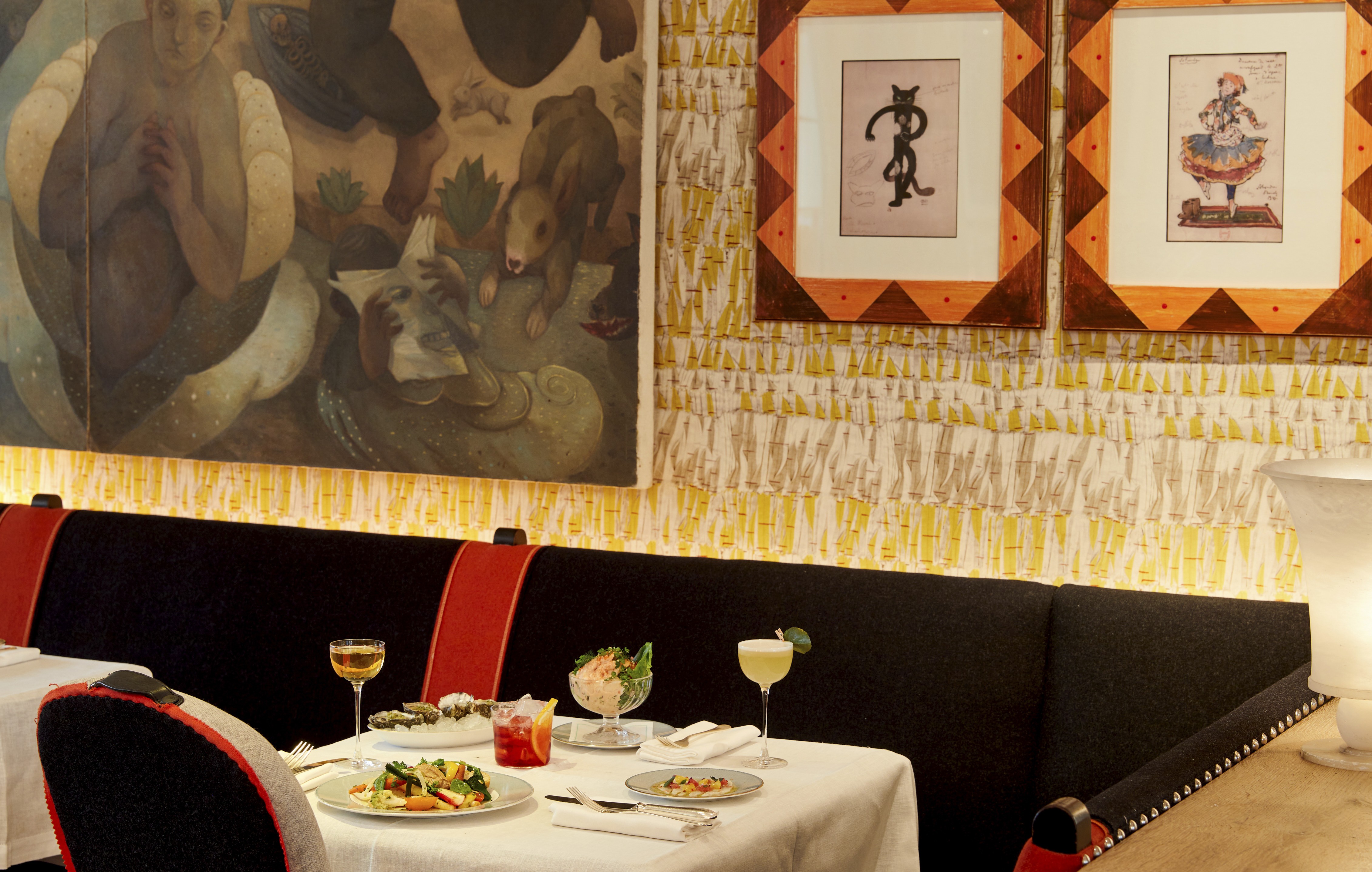 An image to promote the Summer à la carte starters at Ham Yard Bar & Restaurant at Ham Yard Hotel. The image shows a white-linen table set with a selection of starters, including oysters, prawn cocktail, and vibrant salads. Three cocktails are placed alongside the dishes, set against a backdrop of patterned wallpaper and framed artwork