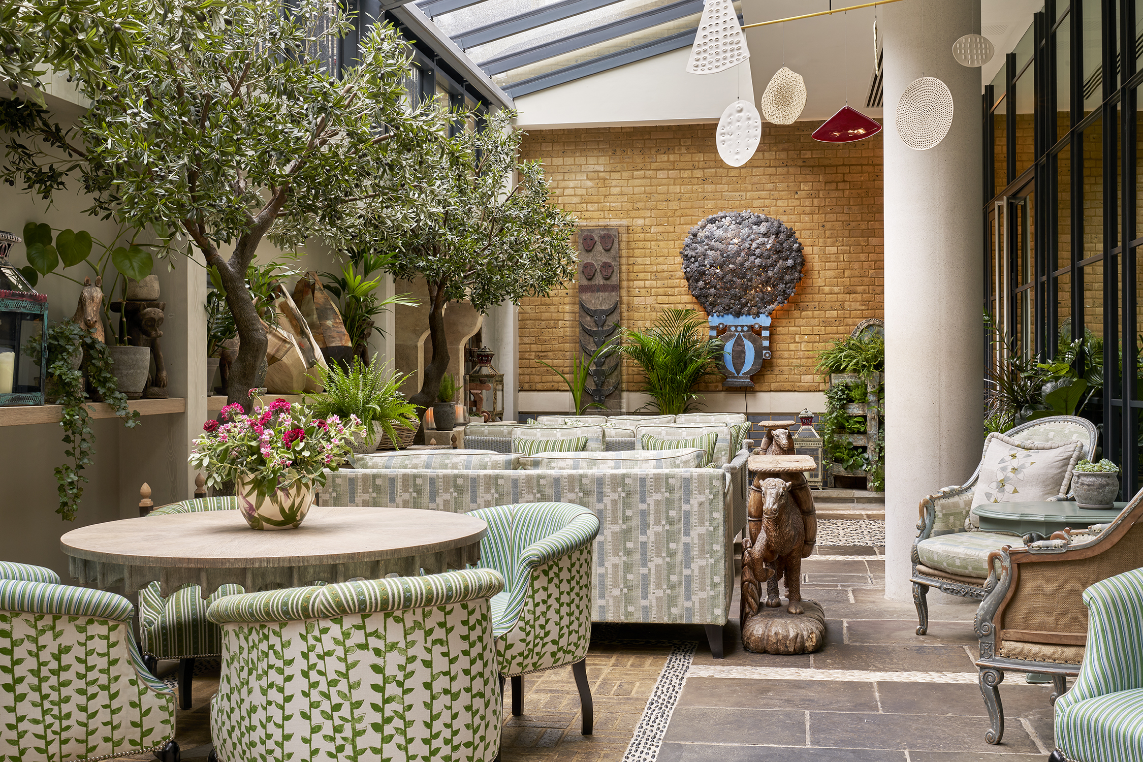 An image of the orangery at the Ham Yard Restaurant, Ham Yard Hotel. The image shows the round table in the foreground of the image with four tub chairs in Little Weed in green around it. Behind that table we see two seating areas both with two sofas facing each other.