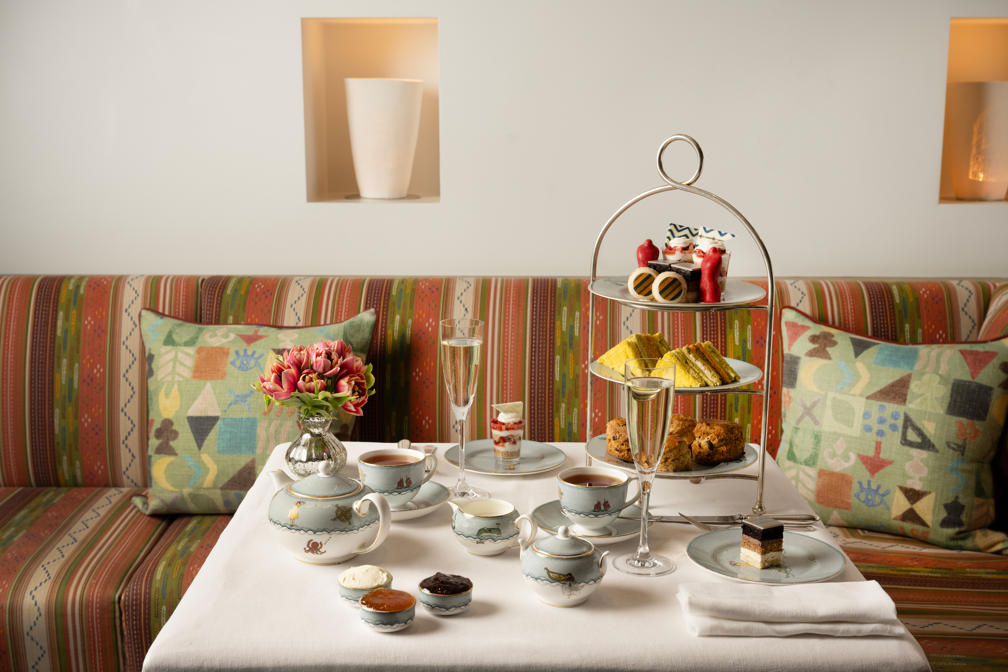 The Kit Kemp Design Stories Afternoon Tea set up at The Whitby Bar. There is a cake stand with scones, cakes decorated with Kit Kemps Famous fabrics, edible mannequins and sandwiches with tea and Champagne