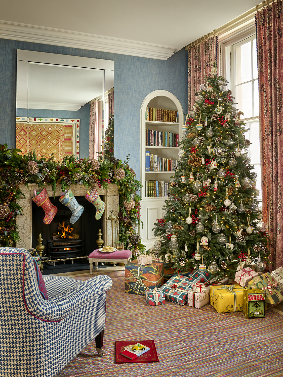 This image shows a beautifully decorated Christmas setting in the townhouse at Haymarket Hotel, featuring a Christmas tree adorned with twinkling lights, red berries, pinecones and ornaments, surrounded by colourful wrapped presents. A roaring fireplace is dressed with festive foliage and stockings, completing the cosy seasonal scene.