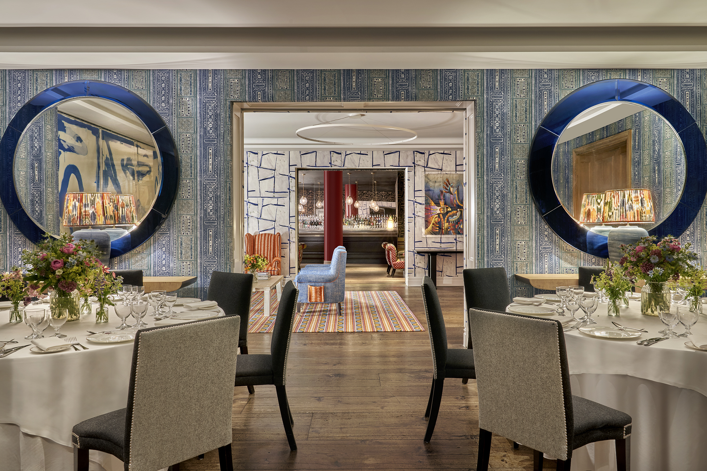 An image of multiple round dining table set up in the Indigo Room, there are white table cloths, floral bouquets in the centre and candles. each place around the table has a placemat, plate, cutlery, glassware and napkins ready for dining. private events room at The Soho Hotel.