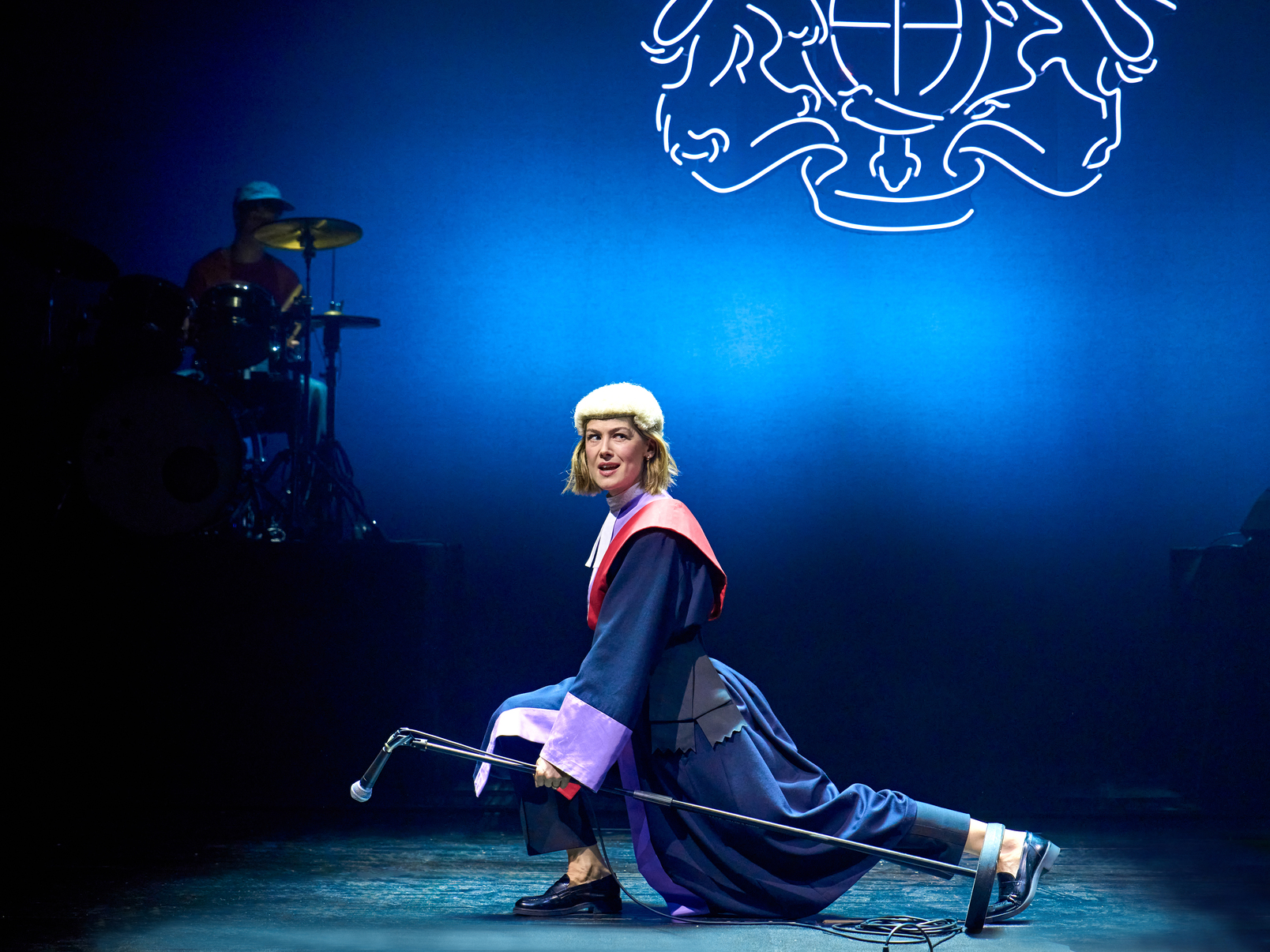 A production shot from Inter Alia. A woman dressed in a judge's wig and robe lunges across the stage holding a microphone stand and looking out in to the audience