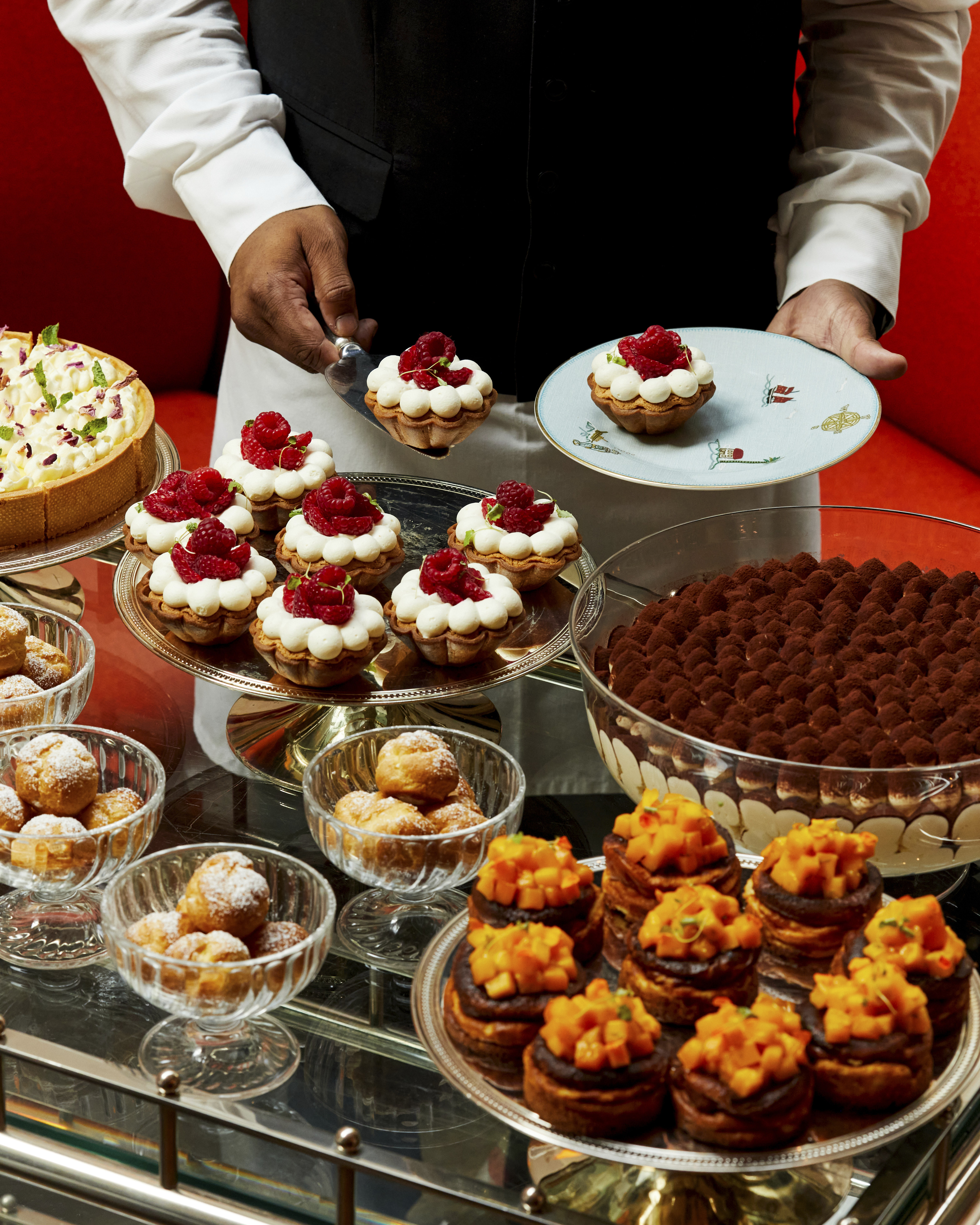 A portrait image taken of The Long Lunch at Ham Yard Bar & Restaurant. The image shows a luxurious dessert trolley laden with an assortment of intricately presented pastries and cakes. A server in formal attire is plating a raspberry tart.