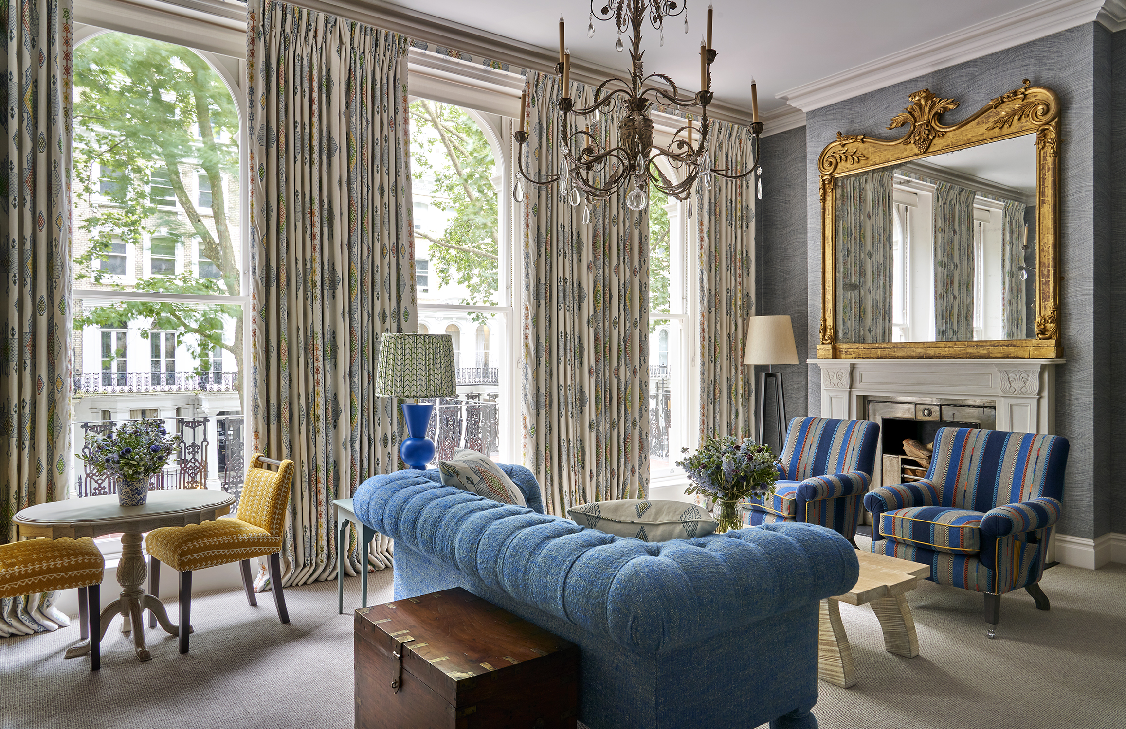 Seating area and fireplace shot of room 101, the Knightsbridge Suite at Knightsbridge Hotel. The image shows a diagonal shot from the bed area across the seating area, showing the blue sofa in the middle of the room facing towards two blue striped armchairs with a low coffee table in the center. A fireplace sits behind the armchair.